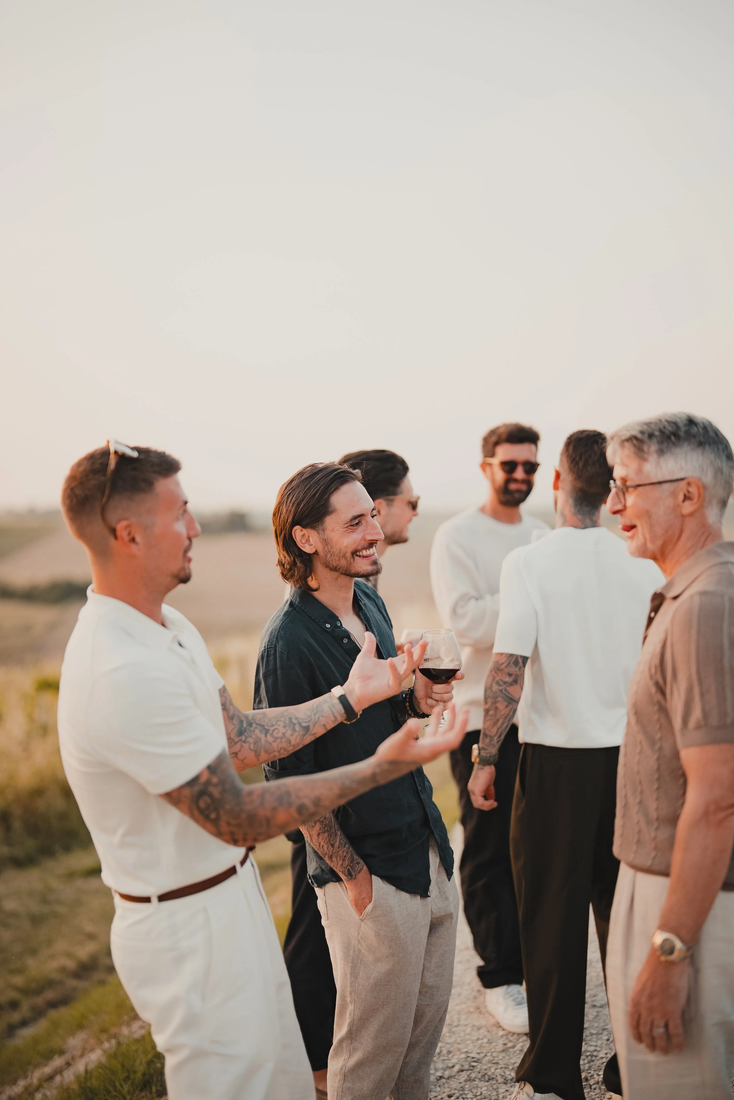 A group of men engaged in conversation outdoors during sunset, some with tattoos and glasses, dressed in smart casual attire.