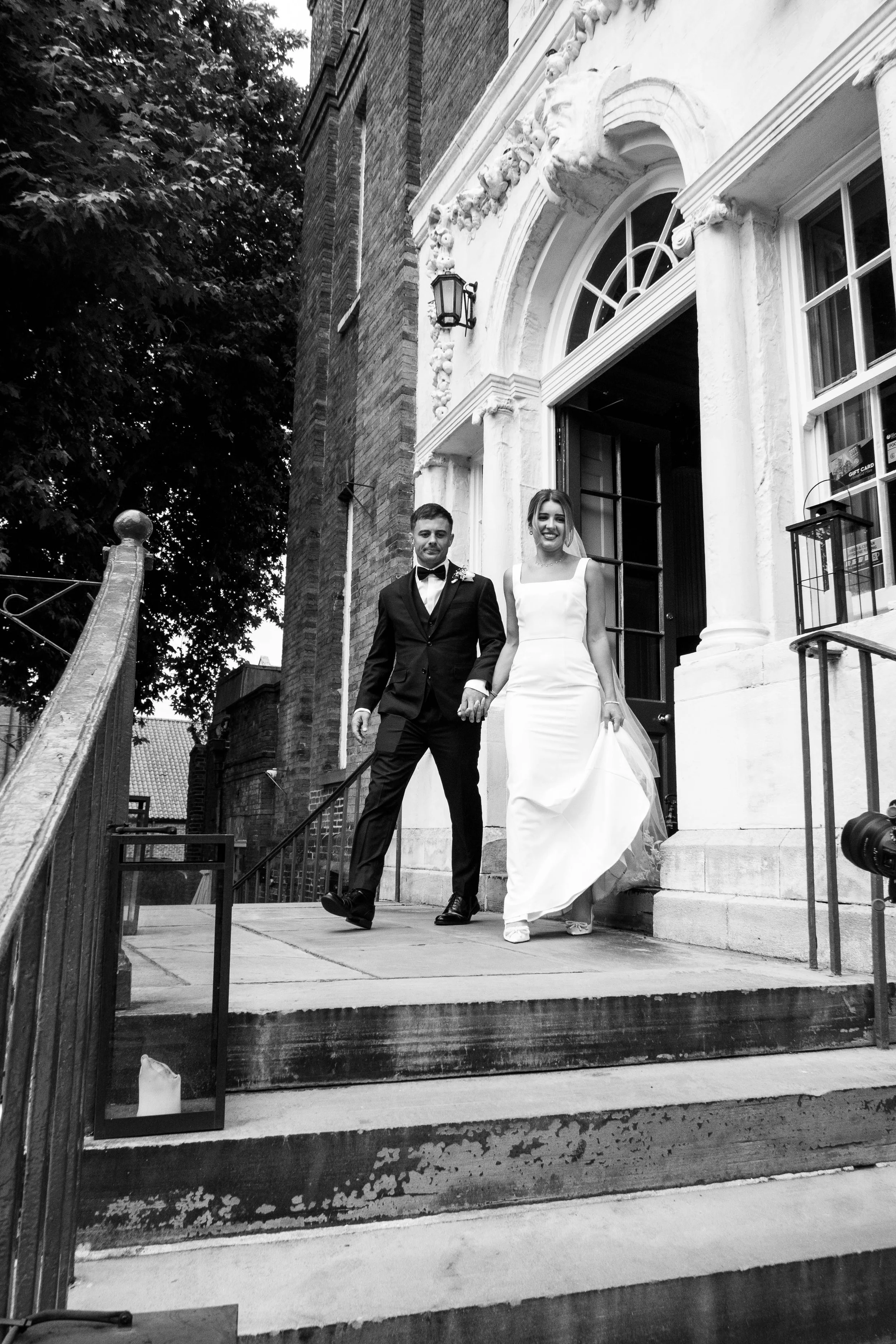 Black and white photo of a newlywed couple descending the stairs of a historic building. The groom is wearing a black tuxedo with a bow tie, and the bride is wearing a white wedding gown. They are holding hands and smiling.