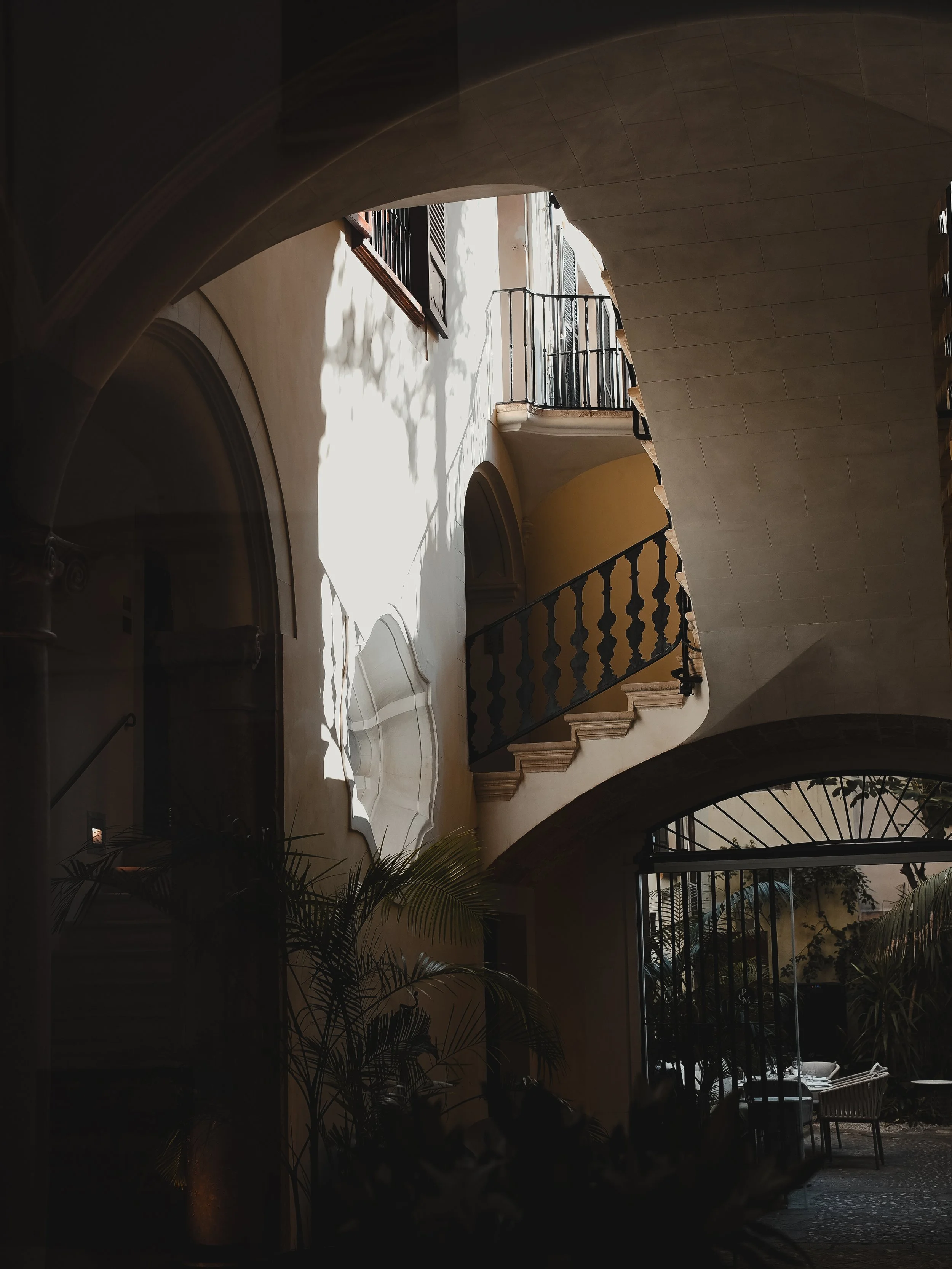Bright sunlight streaming through windows of a hallway, casting shadows. The architecture includes arches, balconies with black metal railings, and lush plants on the ground floor inside a building with beige walls.