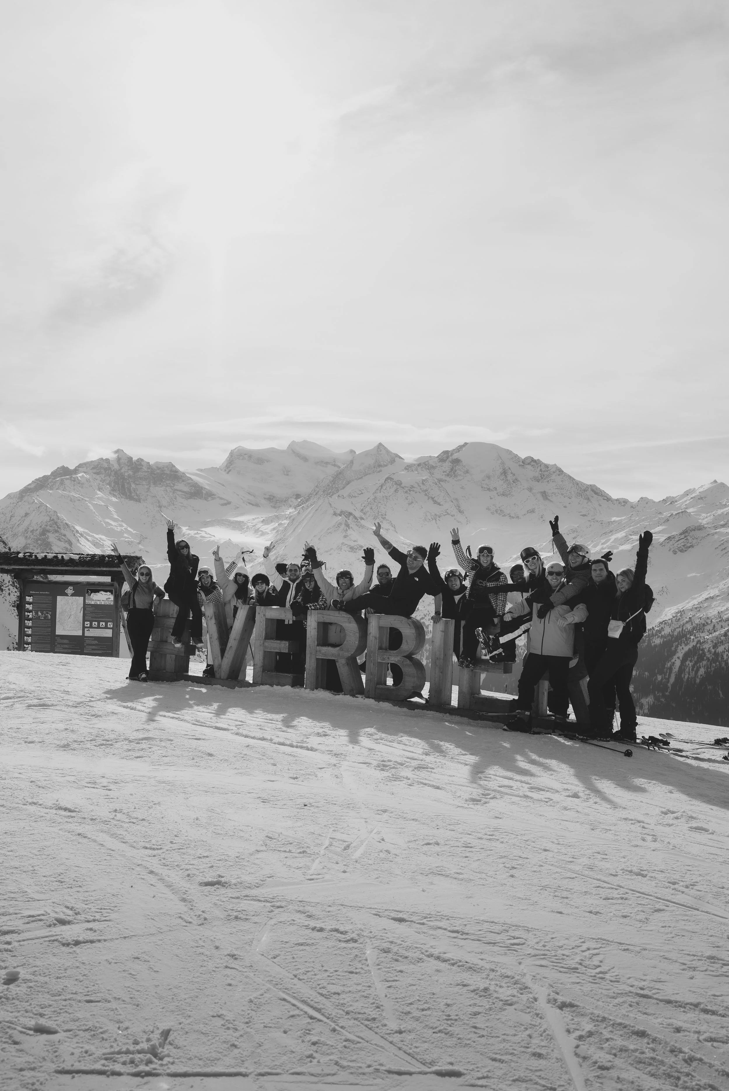 A group of people in ski gear standing behind large wooden letters spelling 'VYRB' on a snowy mountaintop with snow-capped peaks in the background, under a cloudy sky.