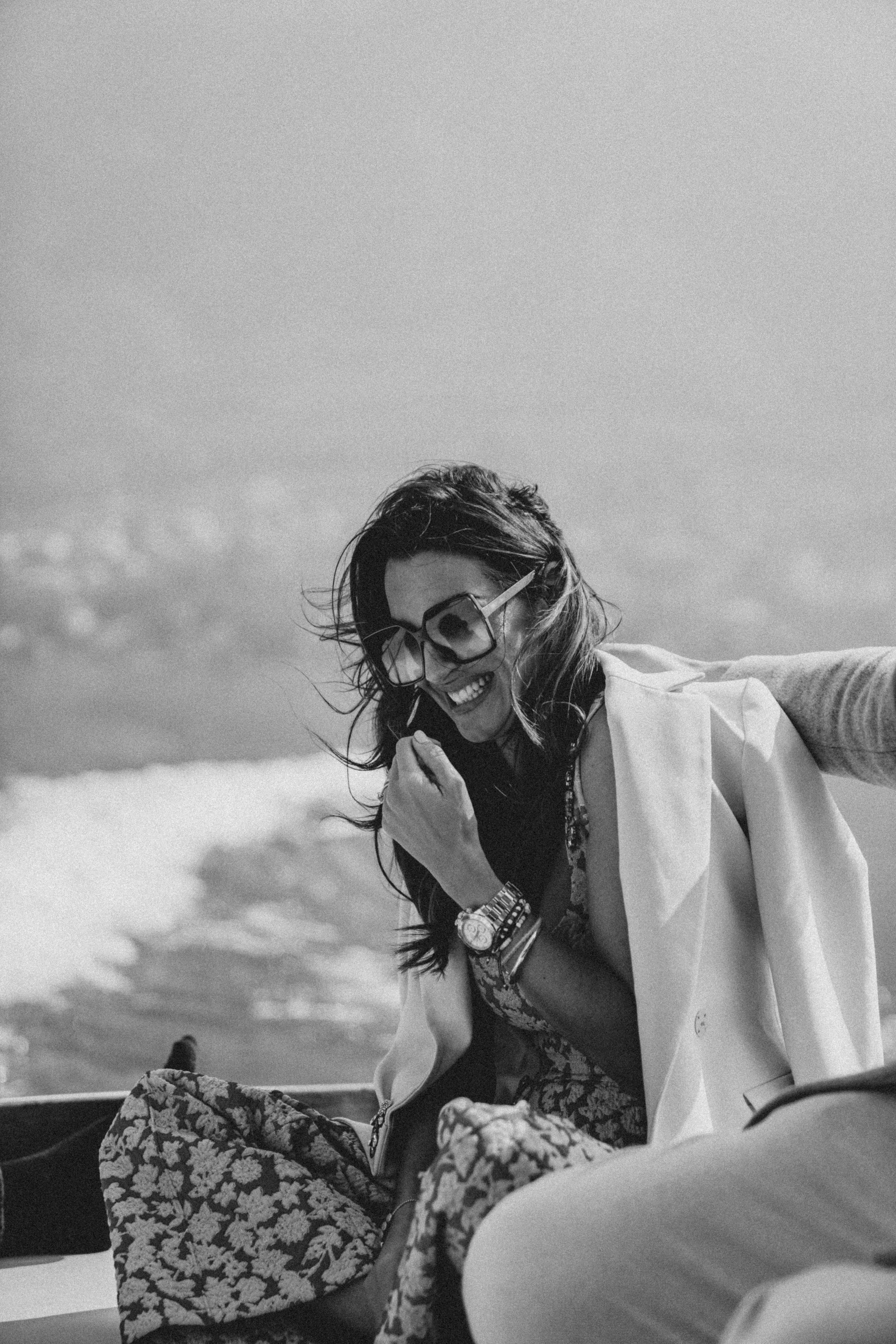 A woman smiling and wearing sunglasses, with her hand near her face, in an outdoor setting with a cloudy sky.