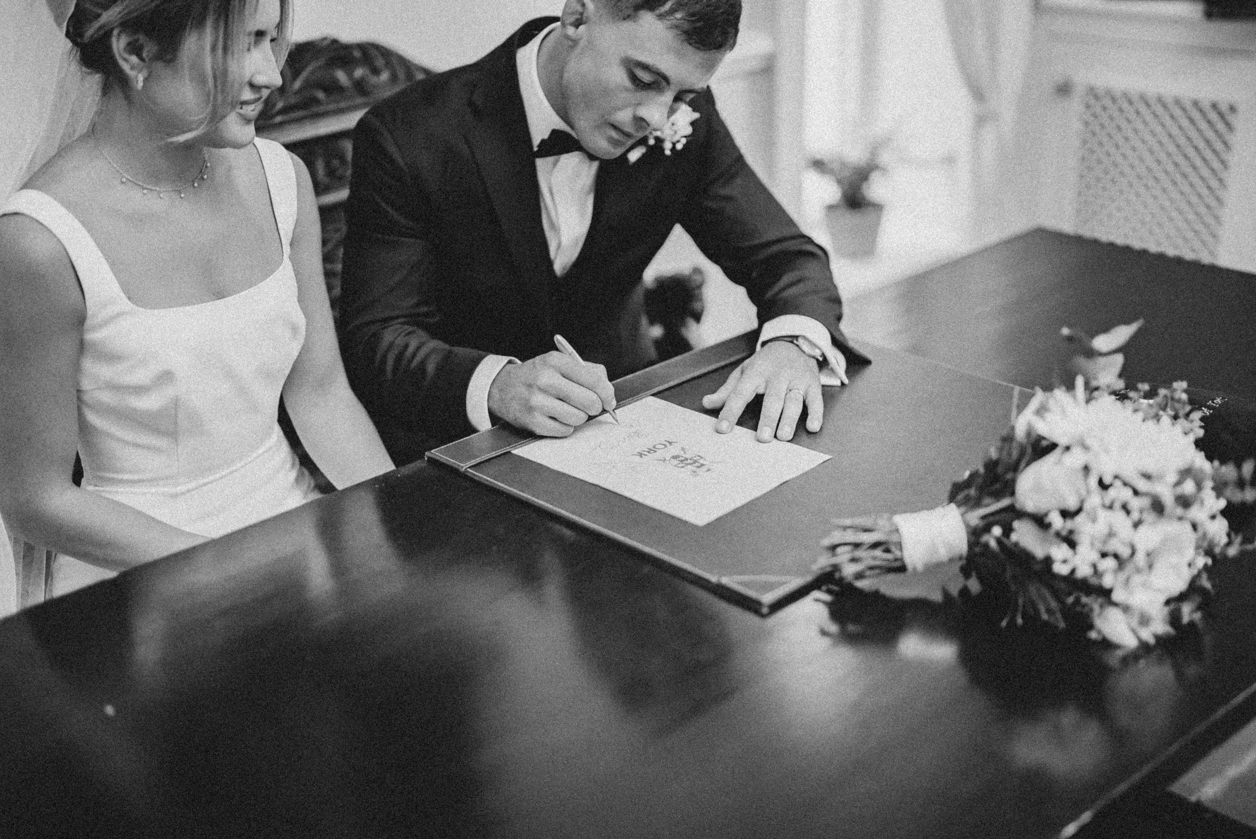 A black-and-white photo of a newlywed couple at their wedding, with the groom signing a marriage document at a table, and the bride watching. There is a bouquet of flowers on the table.