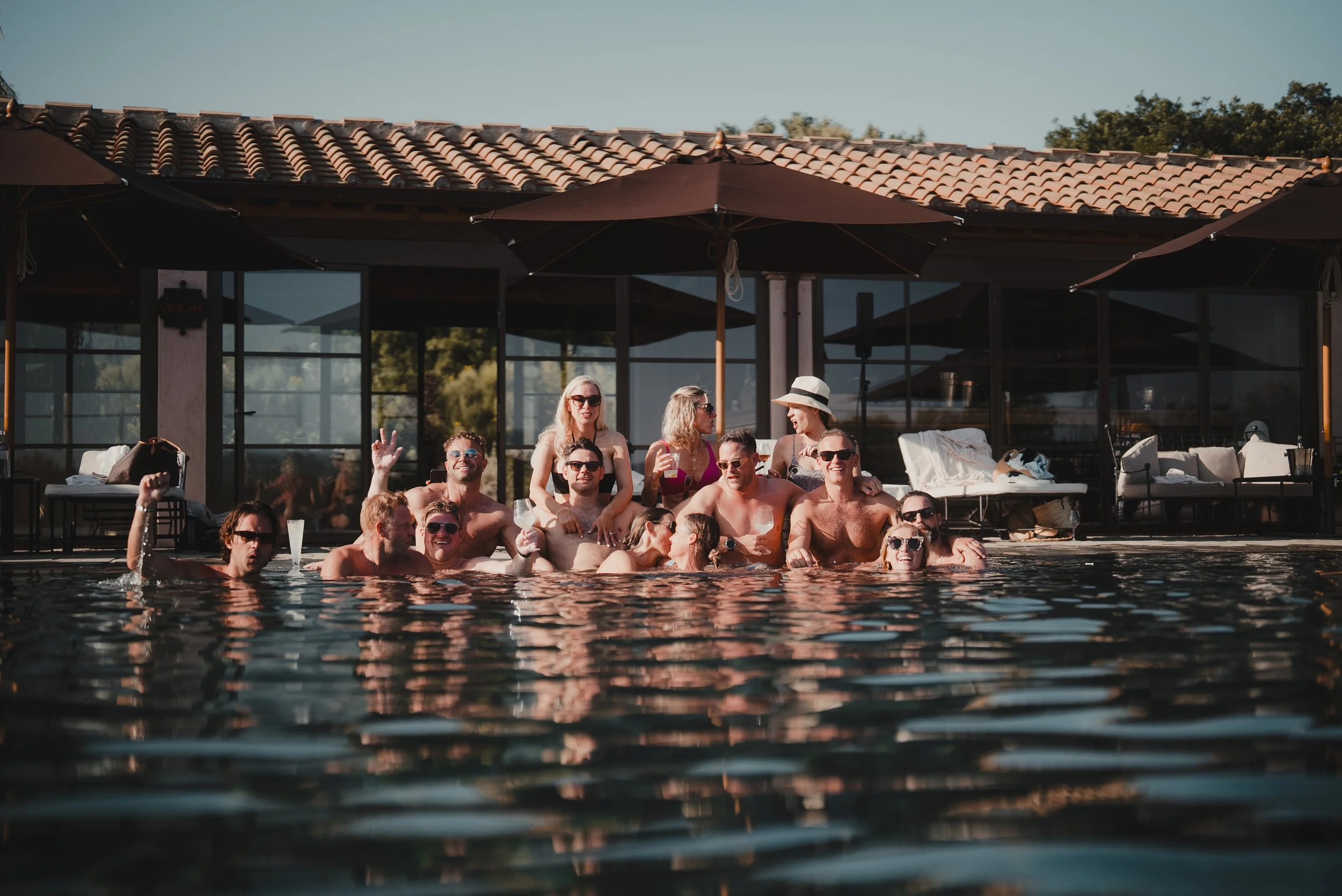 Group of people gathered in a swimming pool during daytime, some wearing sunglasses and holding drinks, with a building featuring a tiled roof and umbrellas in the background.