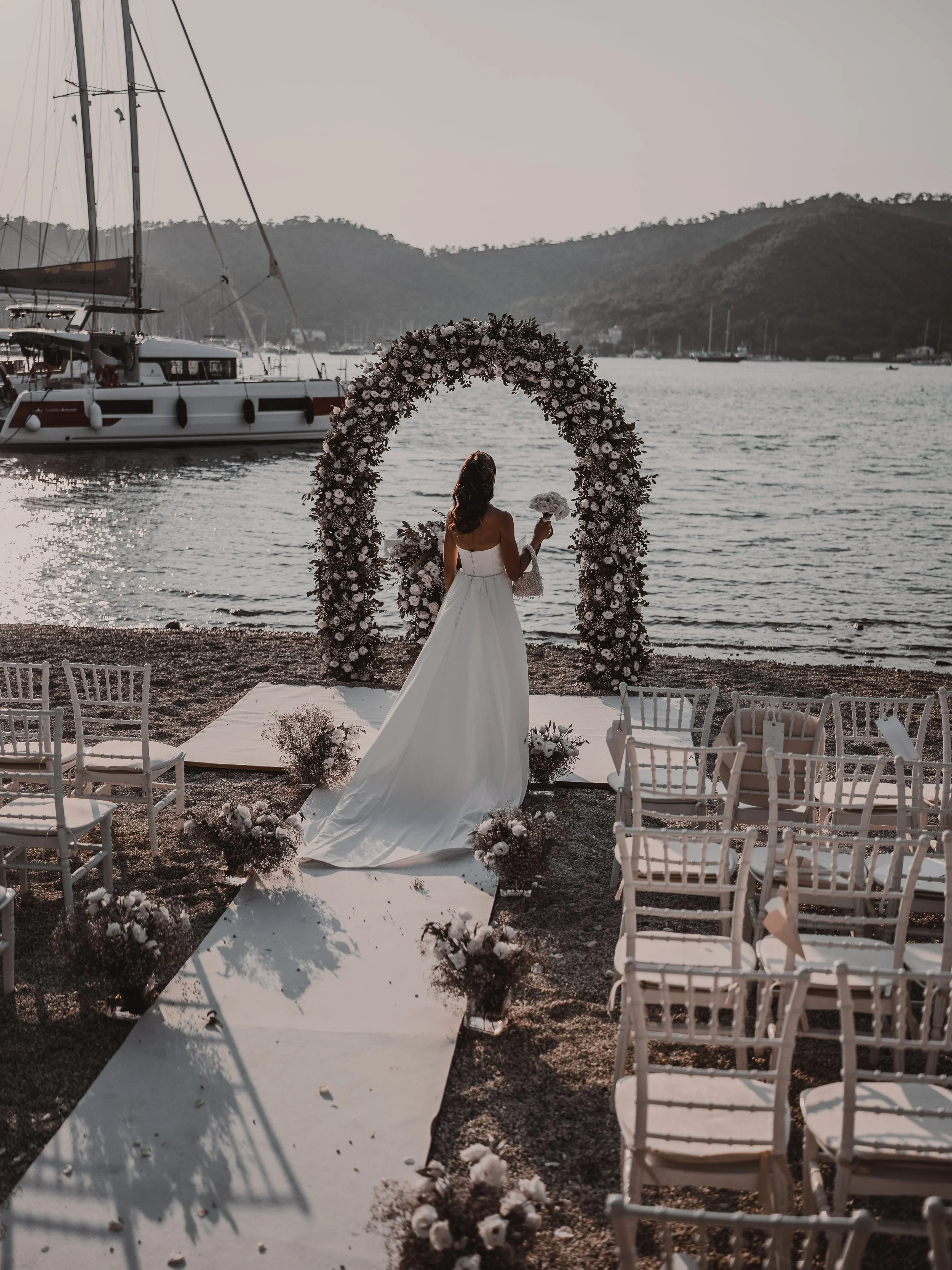A woman in a white wedding dress holding a bouquet stands under a floral arch by the water during a sunset, with a sailboat and mountains in the background.