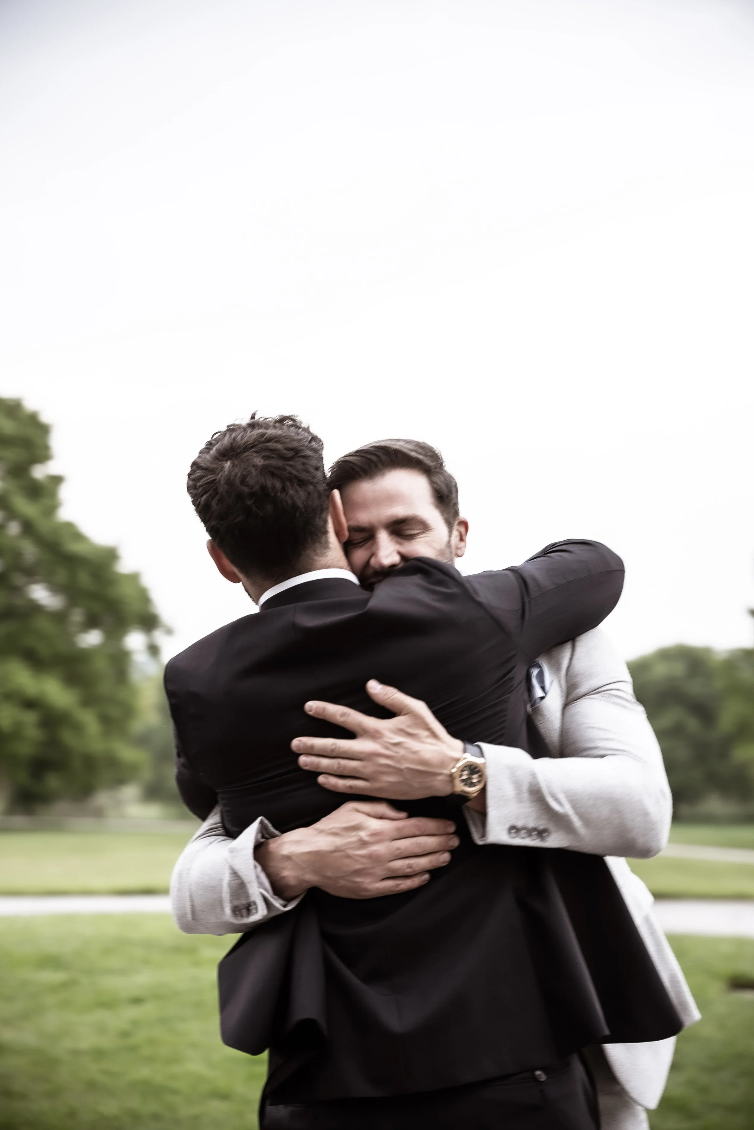 Two men in suits hugging outdoors on a grassy area with trees and a cloudy sky in the background.