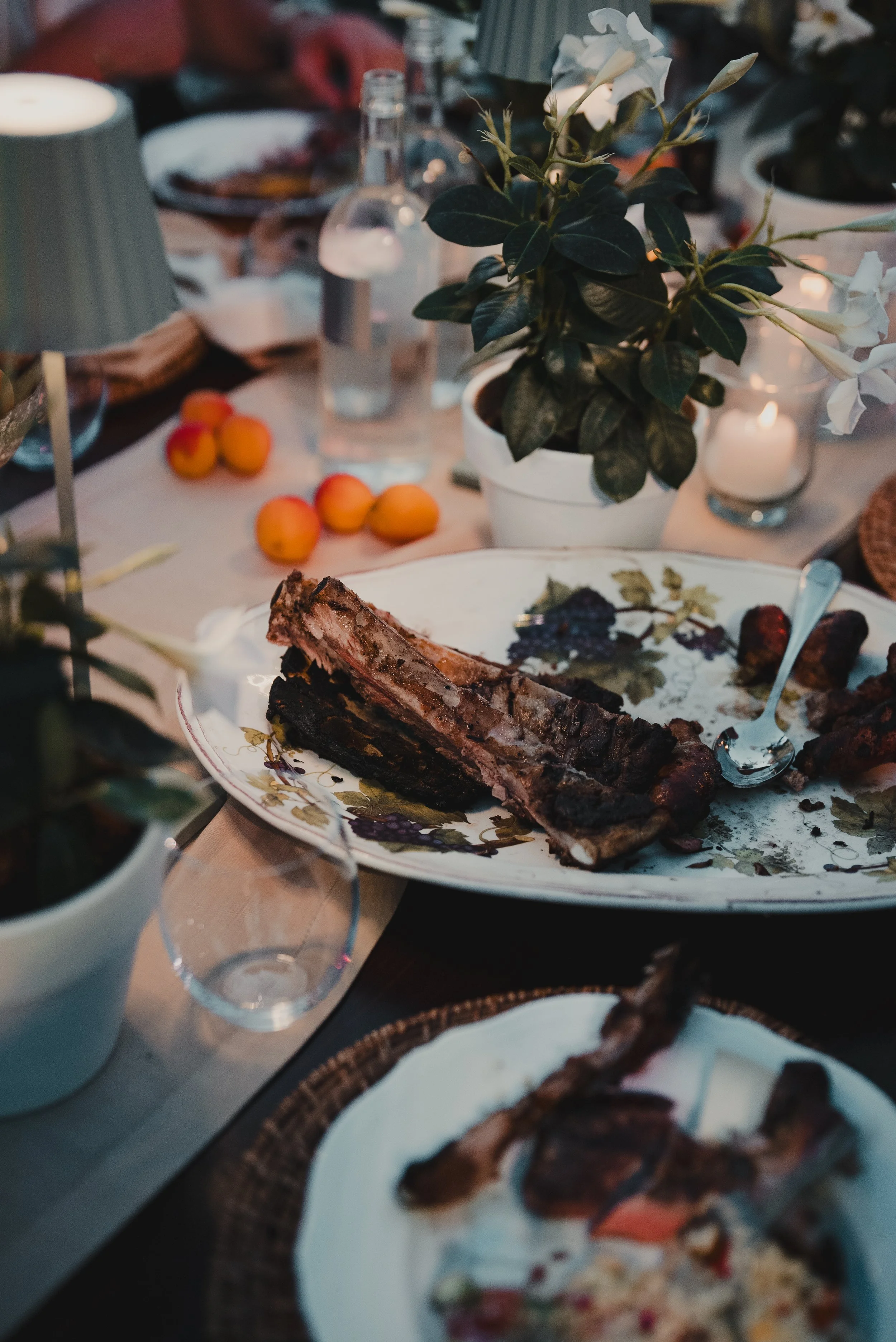 A dining table set for a meal with a centerpiece of a potted white flowering plant, candles, and various plates of food including ribs, fruit, and pizza.