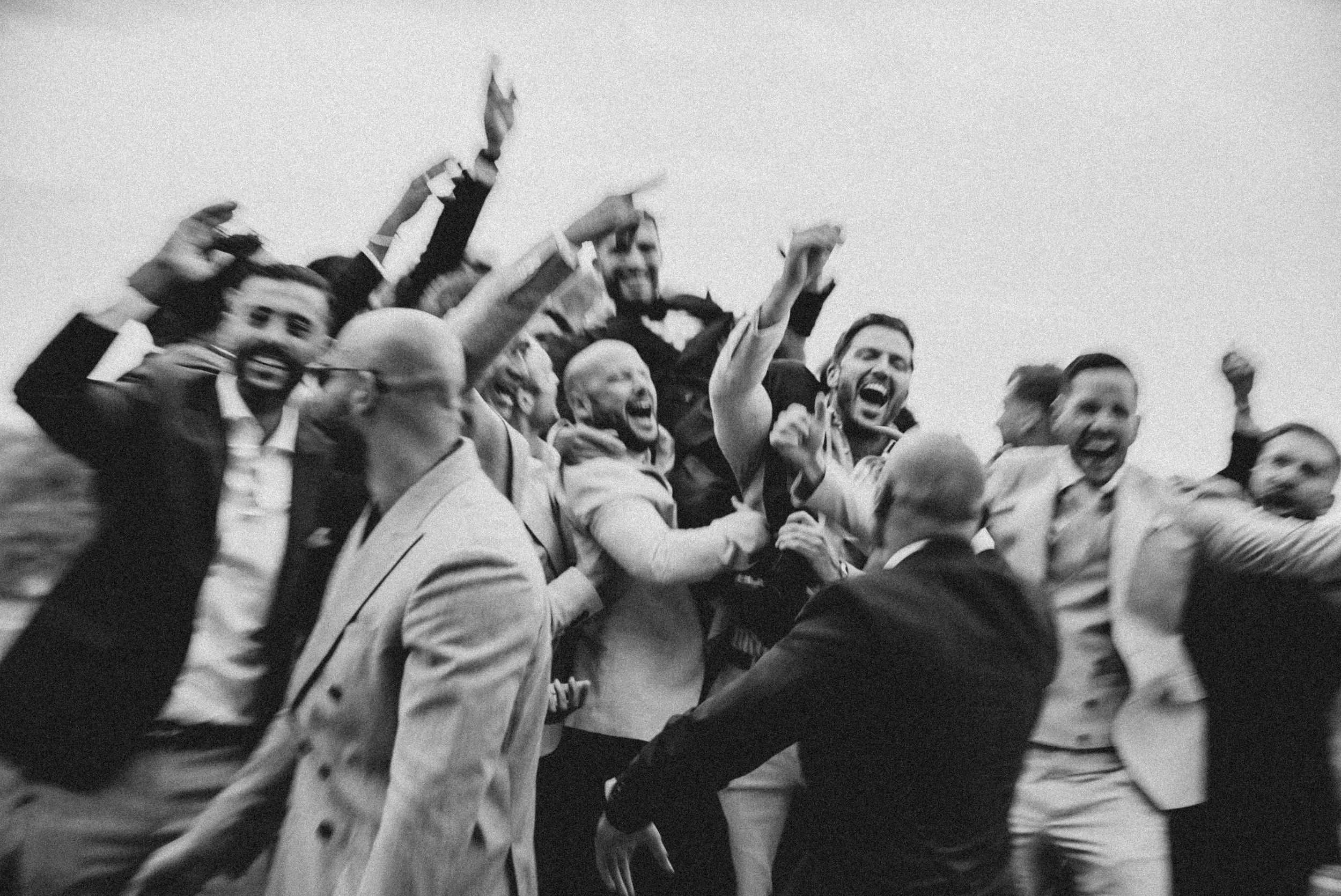 A group of people, mostly men, are celebrating and cheering together outdoors. They are smiling, some with fists raised, and appear to be happy and excited.