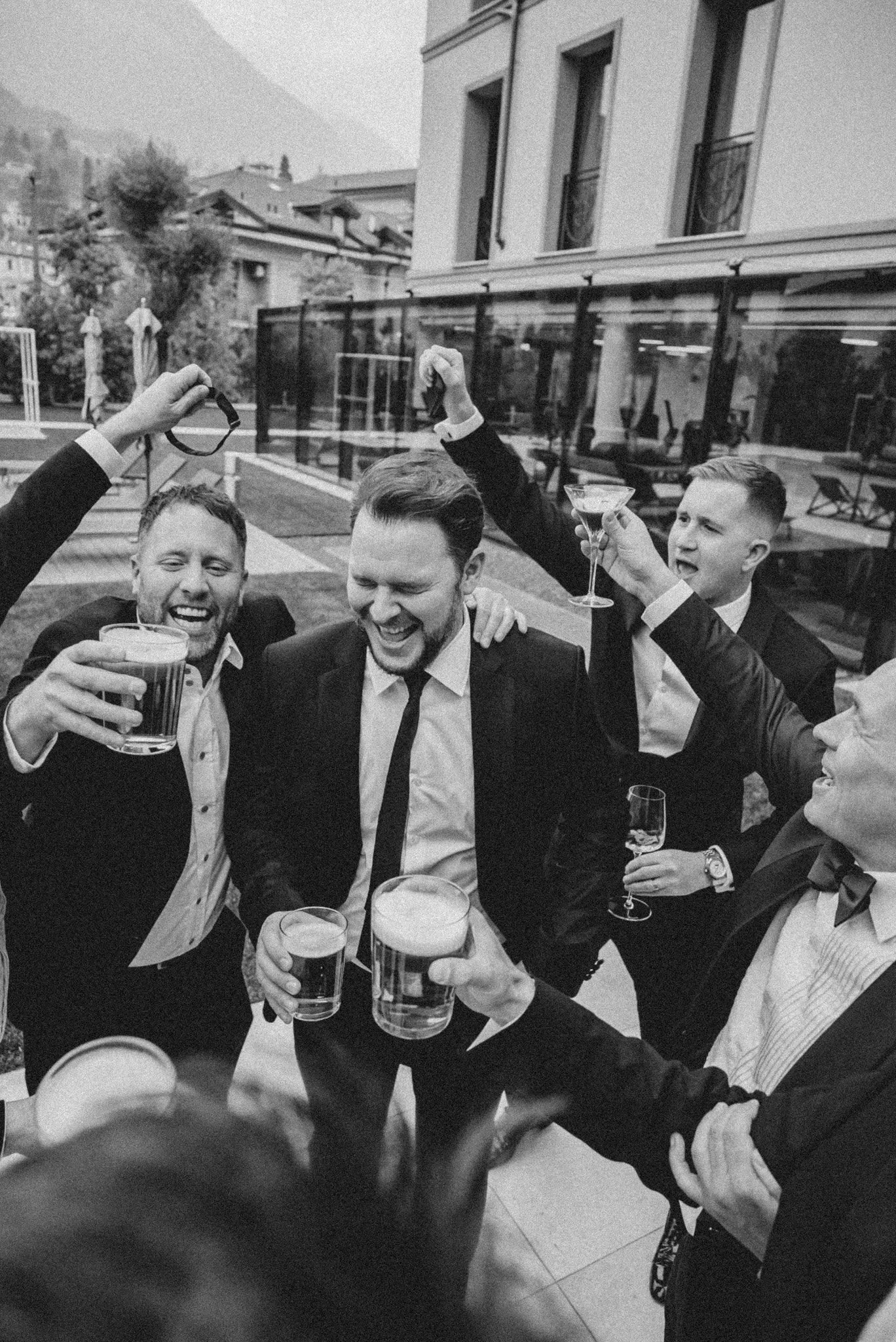A black and white photo of men dressed in suits celebrating at a social gathering, holding drinks, with some smiling and toasting inside a building with large windows and outside visible in the background.