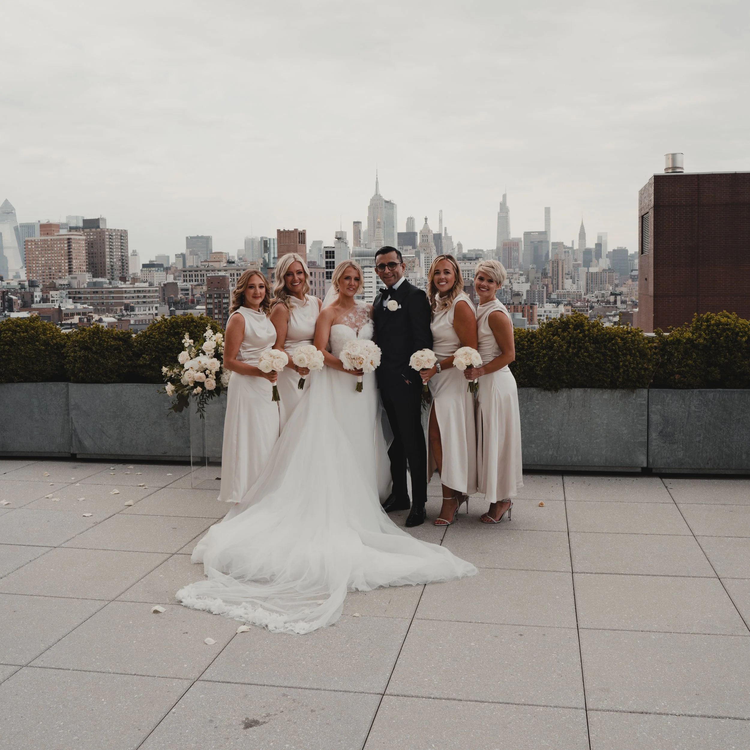 Group of six women and one man in wedding attire standing outdoors on a rooftop with city skyline in the background, smiling for a photo.