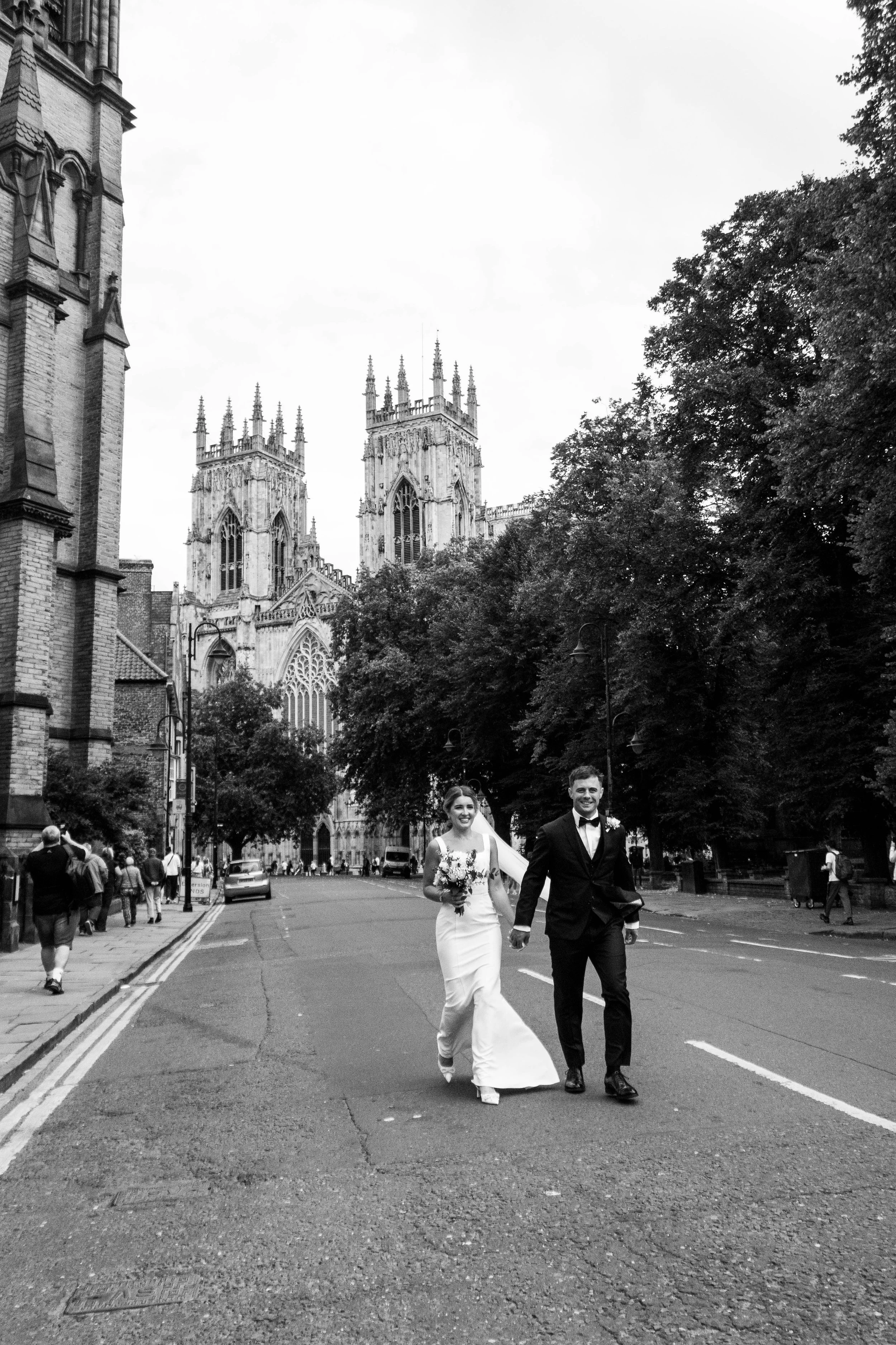 A newlywed couple walks hand in hand down a city street, with the architecture of a historic cathedral and trees in the background, during daylight hours.