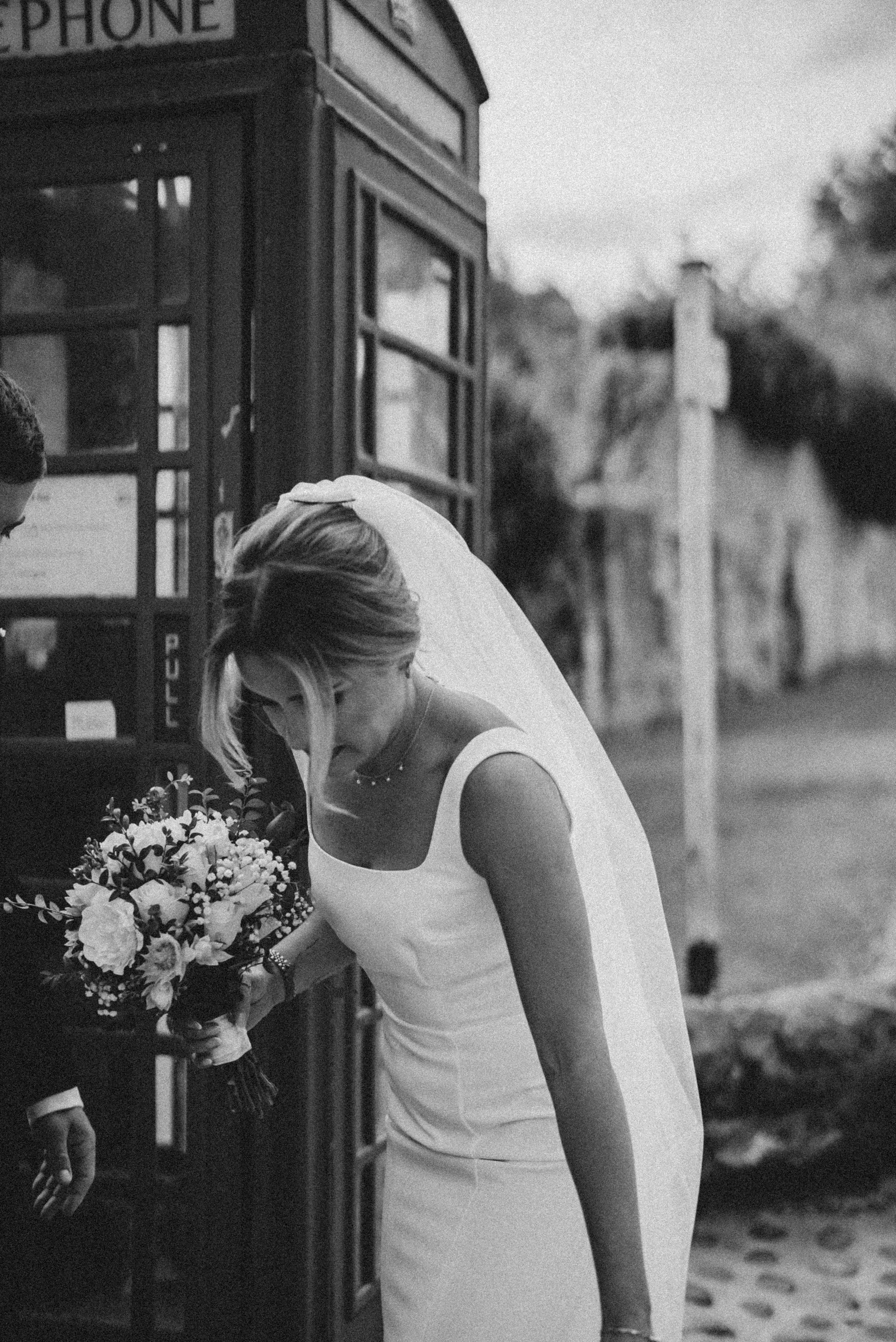 A bride in a white dress and veil holding a large bouquet of flowers, leaning towards a phone booth, with blurred trees and a cloudy sky in the background.