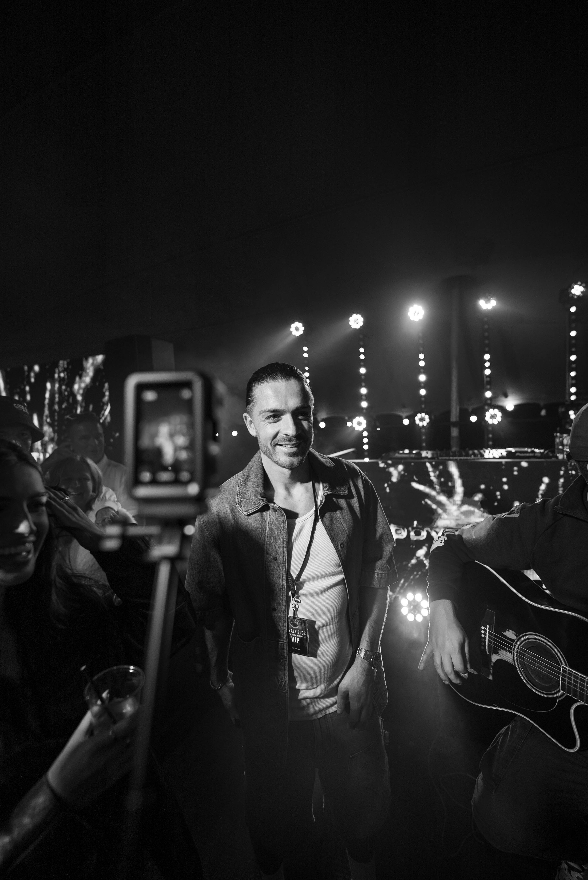 A man with dark hair, dressed in a casual jacket and t-shirt, smiling while surrounded by a group of people. There is a person holding a guitar on his right. They appear to be at a lively event with bright stage lights and a large screen in the backg