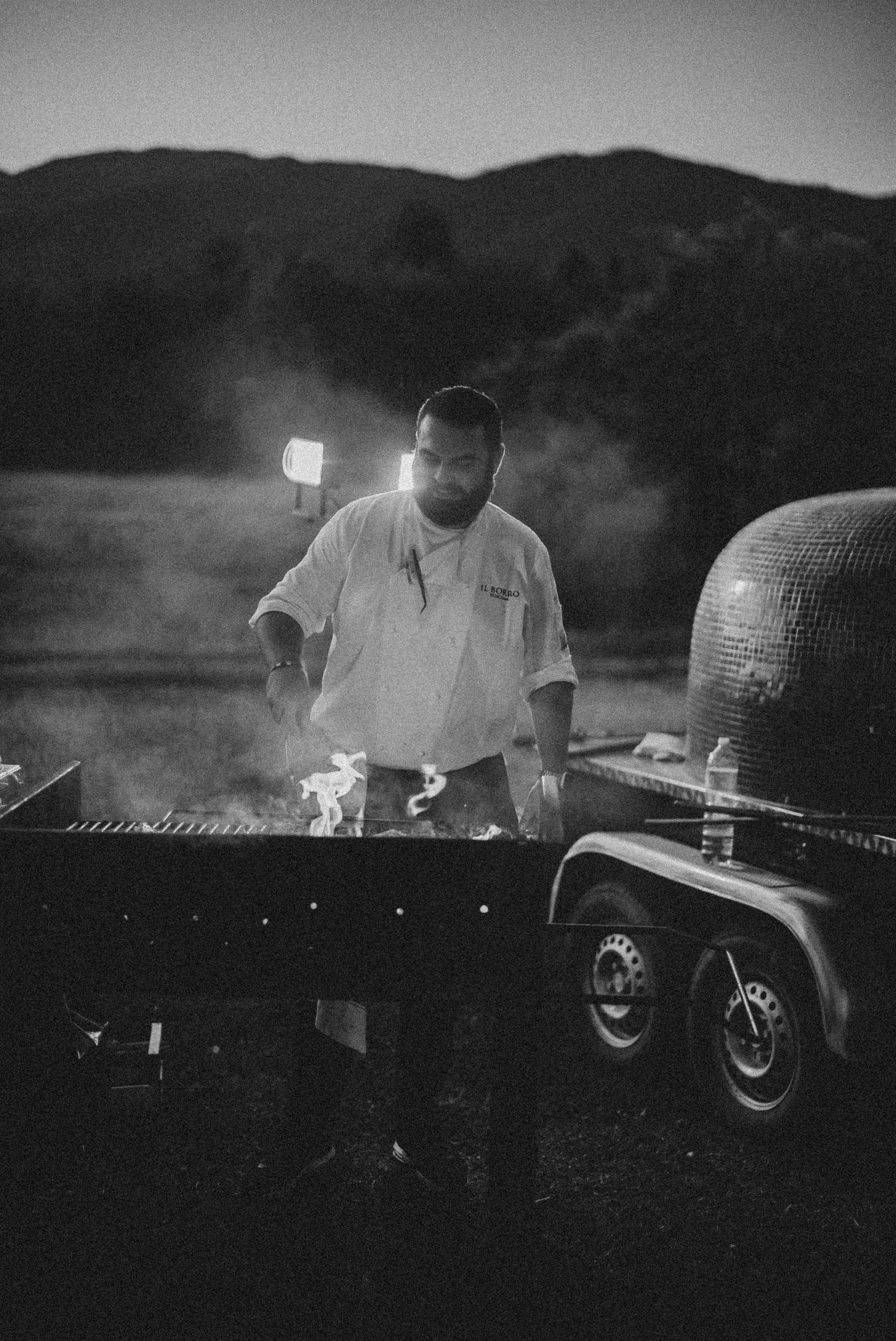 A man with a beard and short hair cooking at an outdoor grill during the evening or night, with mountains in the background and two bright lights illuminating the scene.