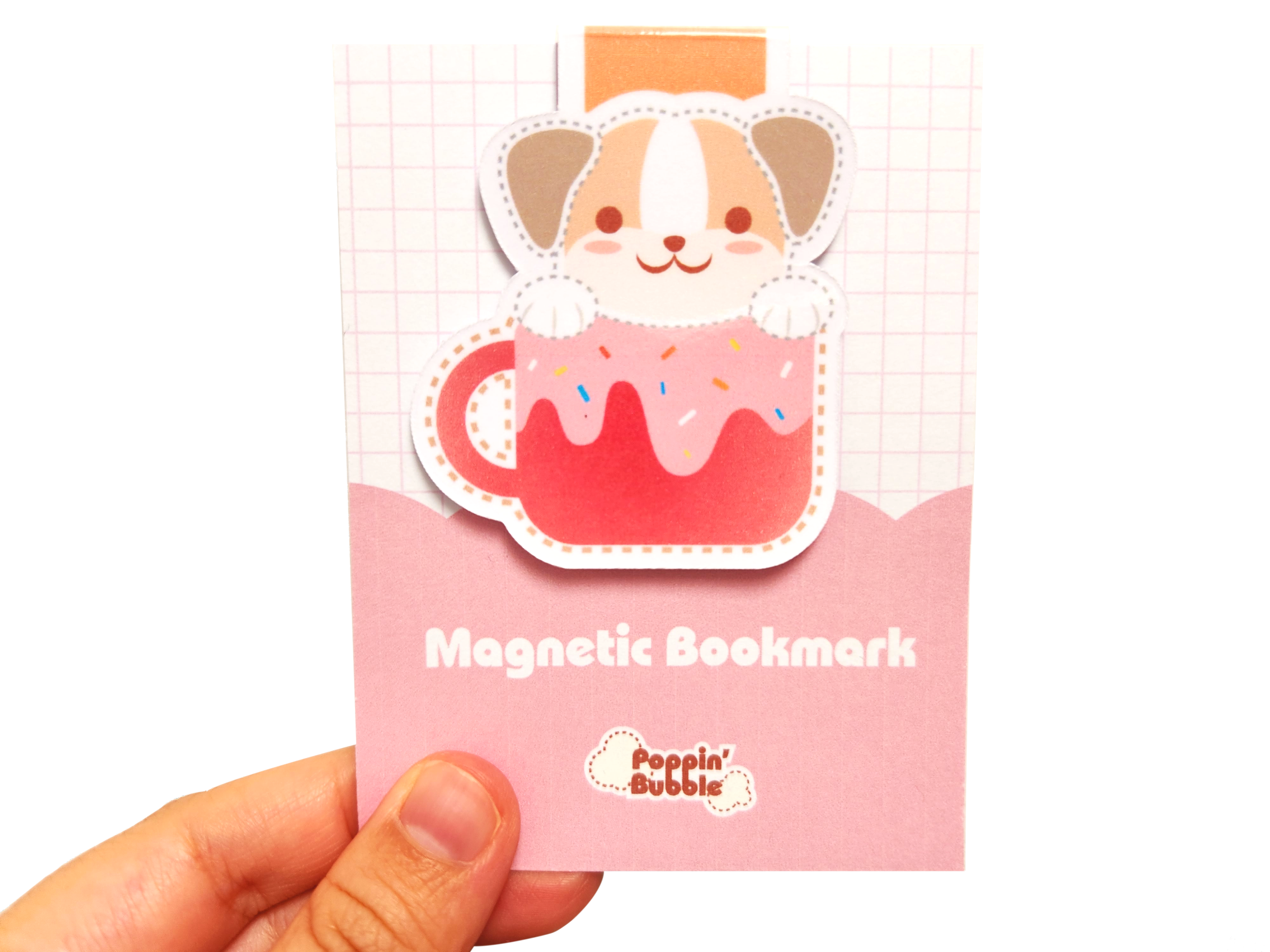 A hand holding a pink card with a cartoon dog in a pink mug with sprinkles. The card says 'Magnetic Bookmark' and 'Poppin' Bubble'.