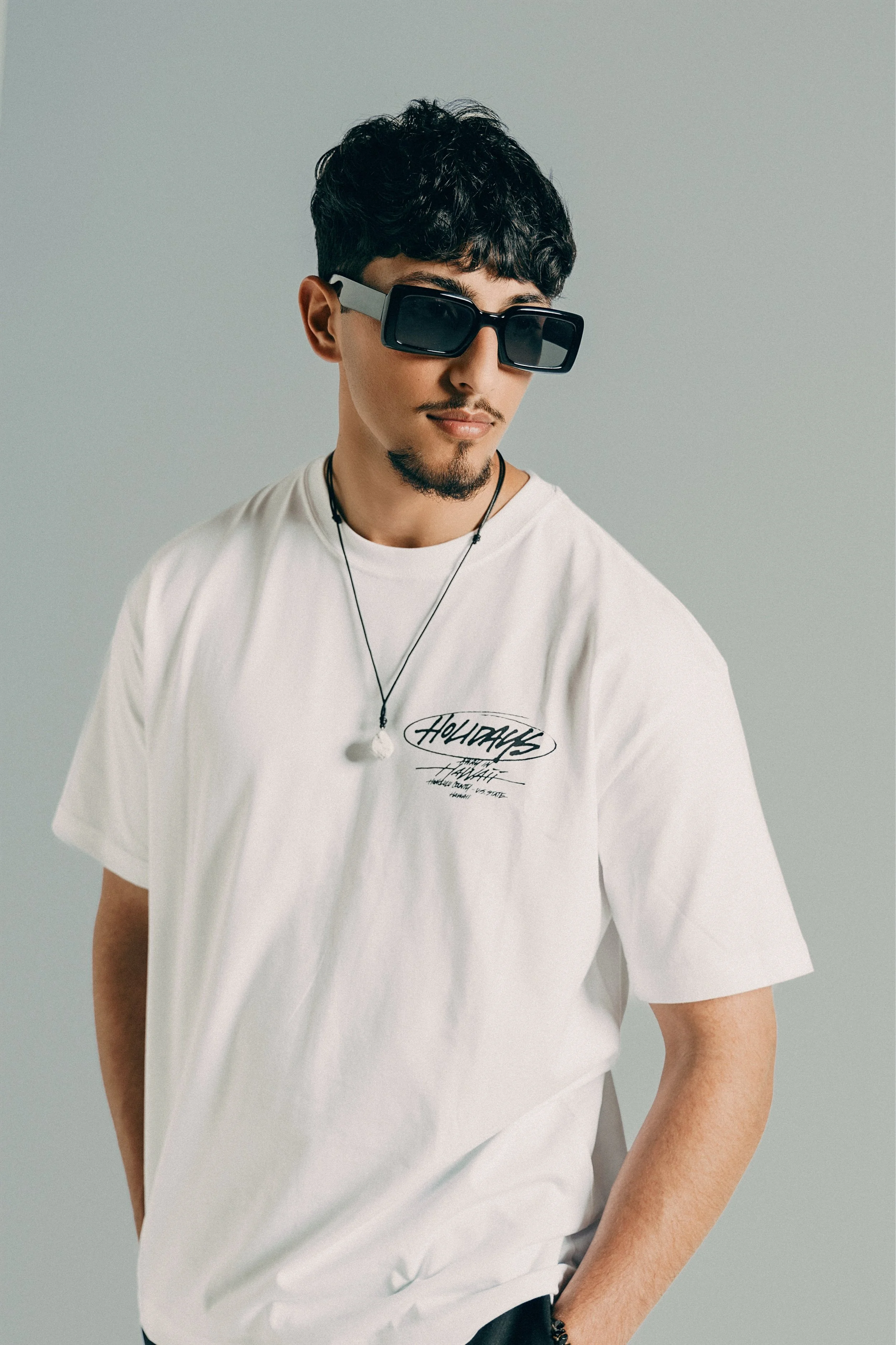 Young man with dark curly hair, wearing large black sunglasses, a white t-shirt with black graphics, and a necklace, posing against a plain light gray background.