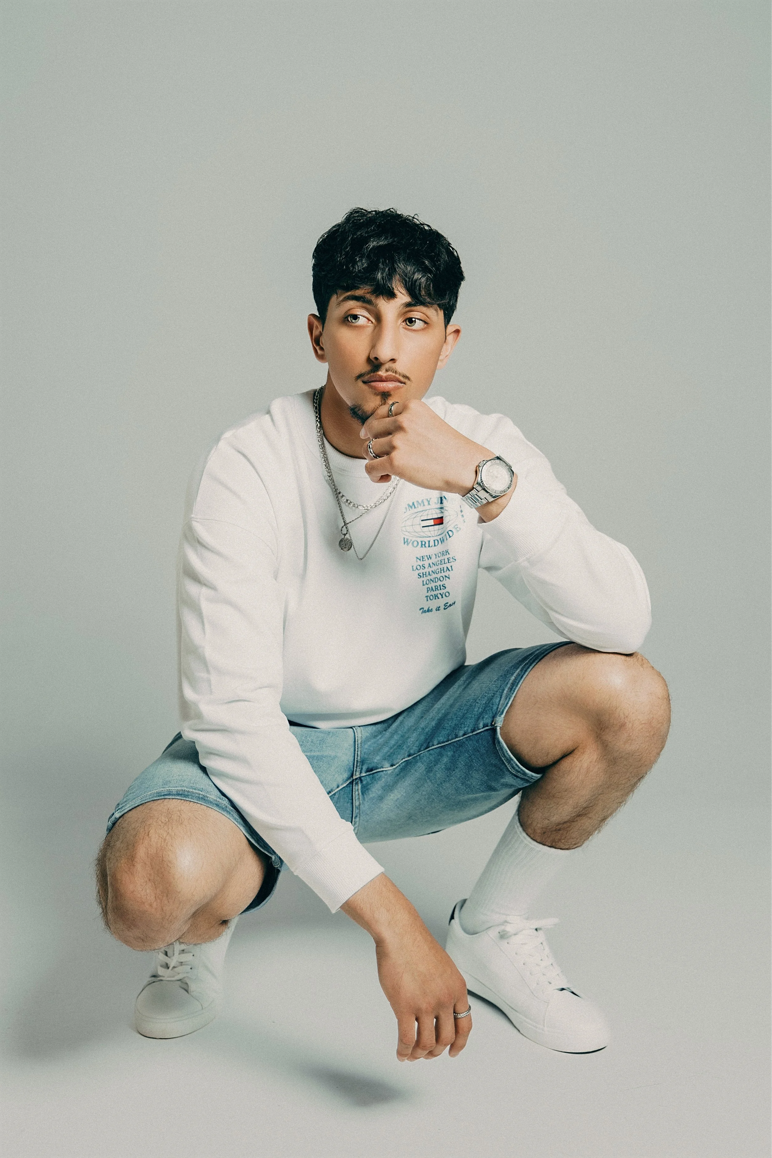 A young man crouching in a studio, wearing a white long-sleeve shirt, denim shorts, white sneakers, and accessories, with a neutral gray background.