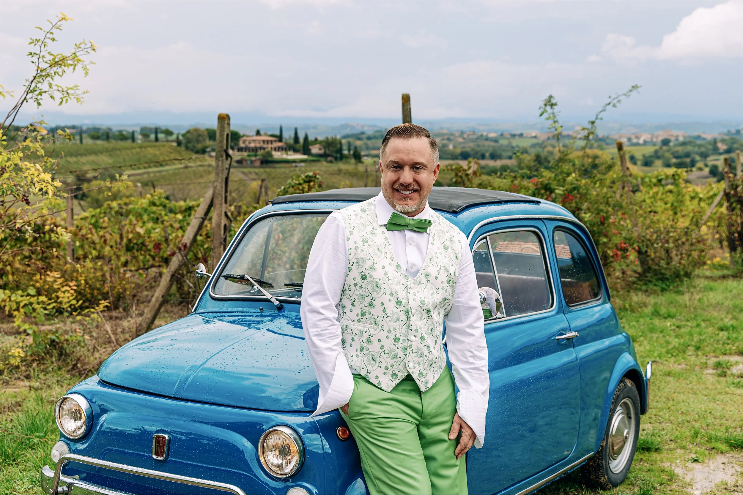A man in a white shirt with a green bow tie and green pants, standing next to a small blue vintage car in a vineyard landscape with rolling hills and scattered houses in the background.