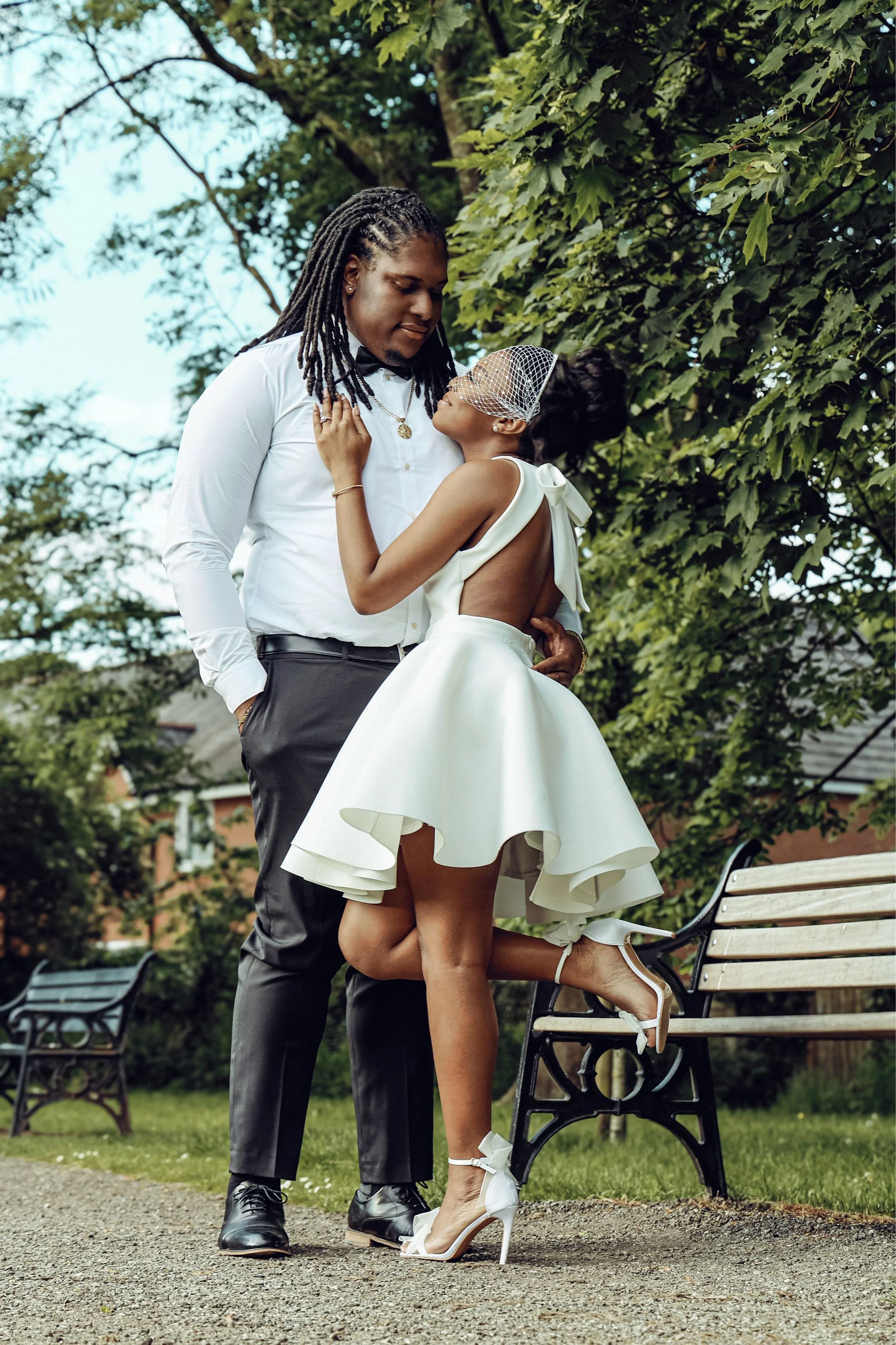 A bride and groom standing close together outdoors, with the bride lifting one leg onto a bench. The bride wears a white dress with a bow on the back, high heels, and a facial net. The groom wears a white shirt with a bowtie and dark trousers. They a