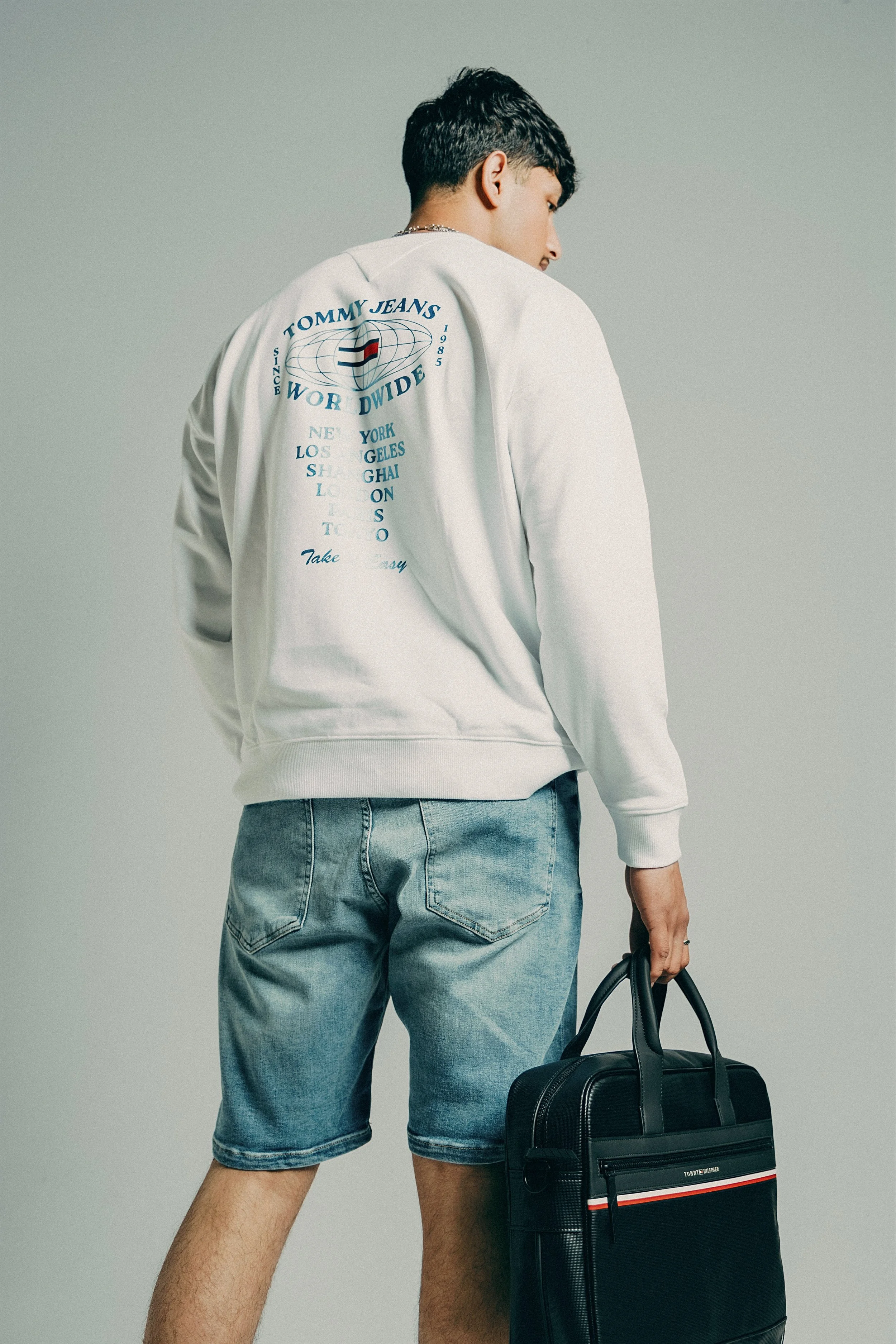 A person wearing a white sweatshirt with blue text and logos on the back, light blue denim shorts, holding a black tote bag with red, white, and black stripe, standing against a plain gray background