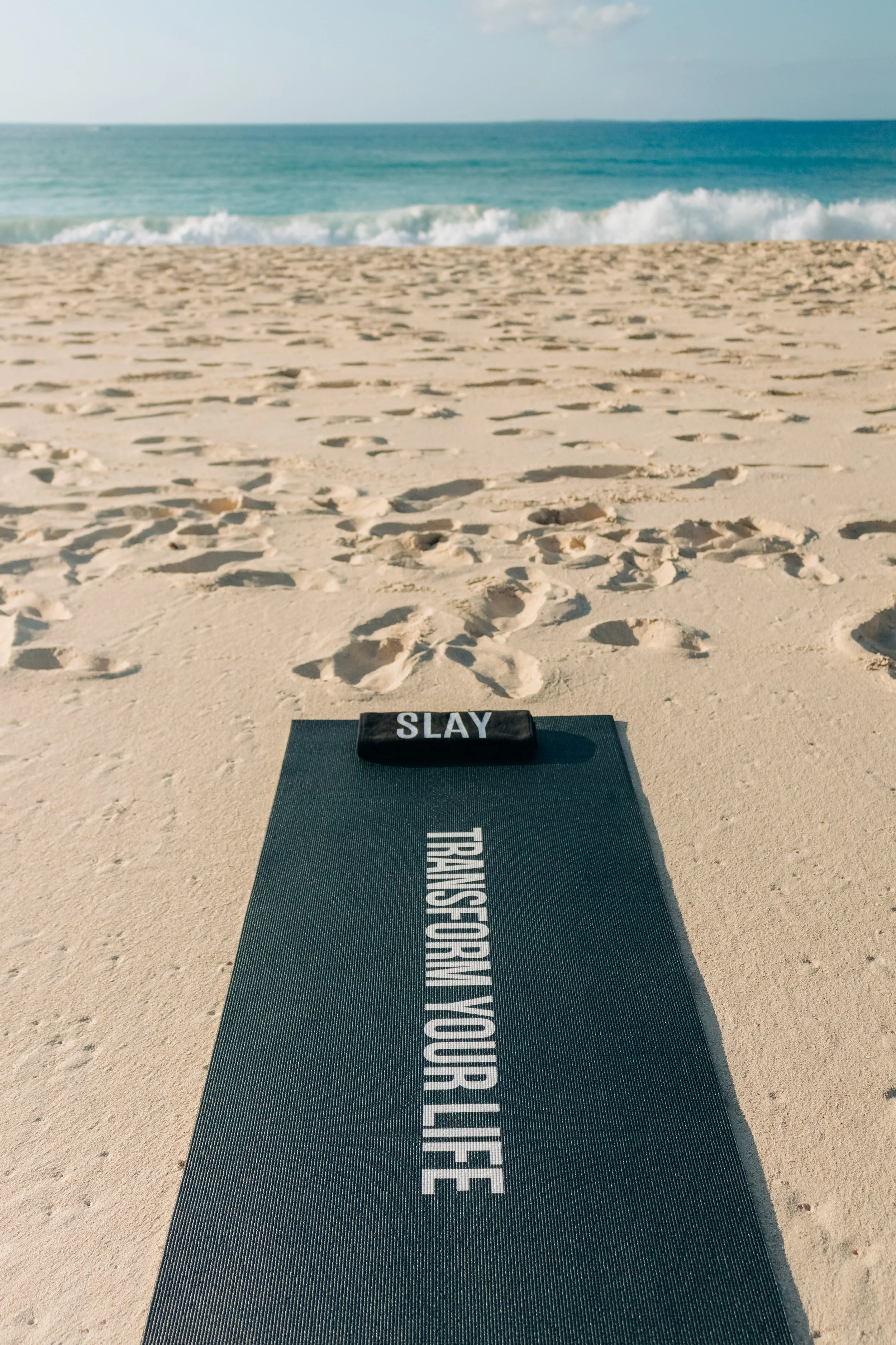 A yoga mat on sandy beach with the words 'TRANSFORM YOUR LIFE' and a small black towel with 'SLAY' written on it, overlooking the ocean with gentle waves and a clear sky.