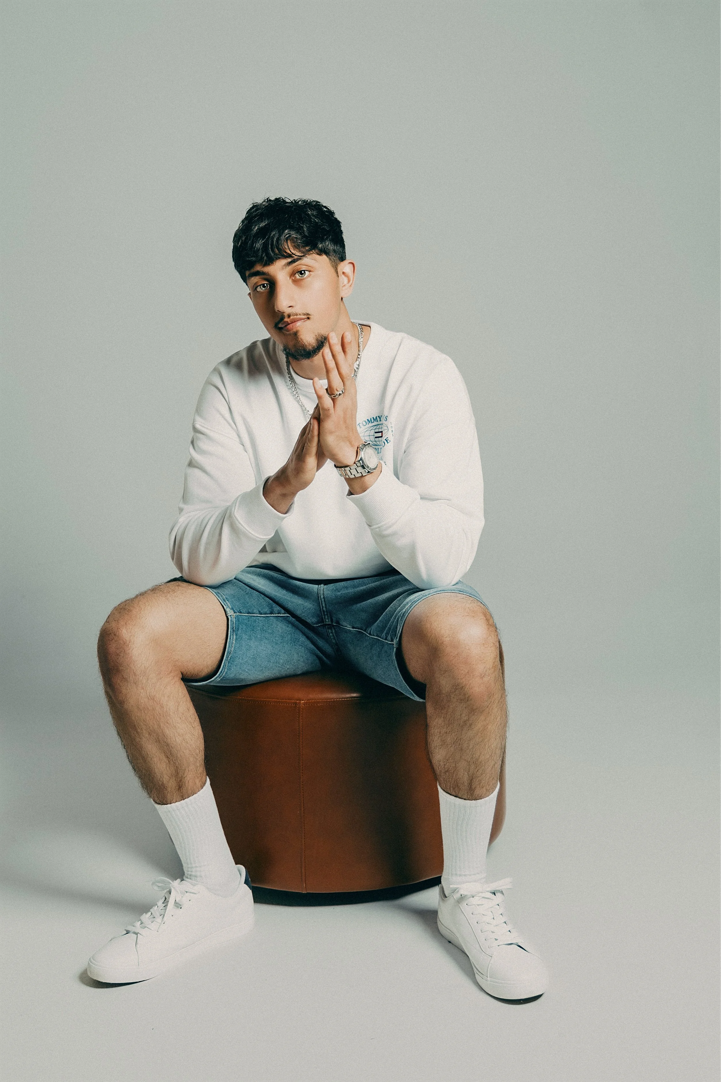 A young man sitting on a brown ottoman in front of a plain, light background. He is wearing a white sweatshirt, denim shorts, white sneakers, and white socks. He has dark hair, a trimmed beard, and is holding his hands together in a prayer-like posit