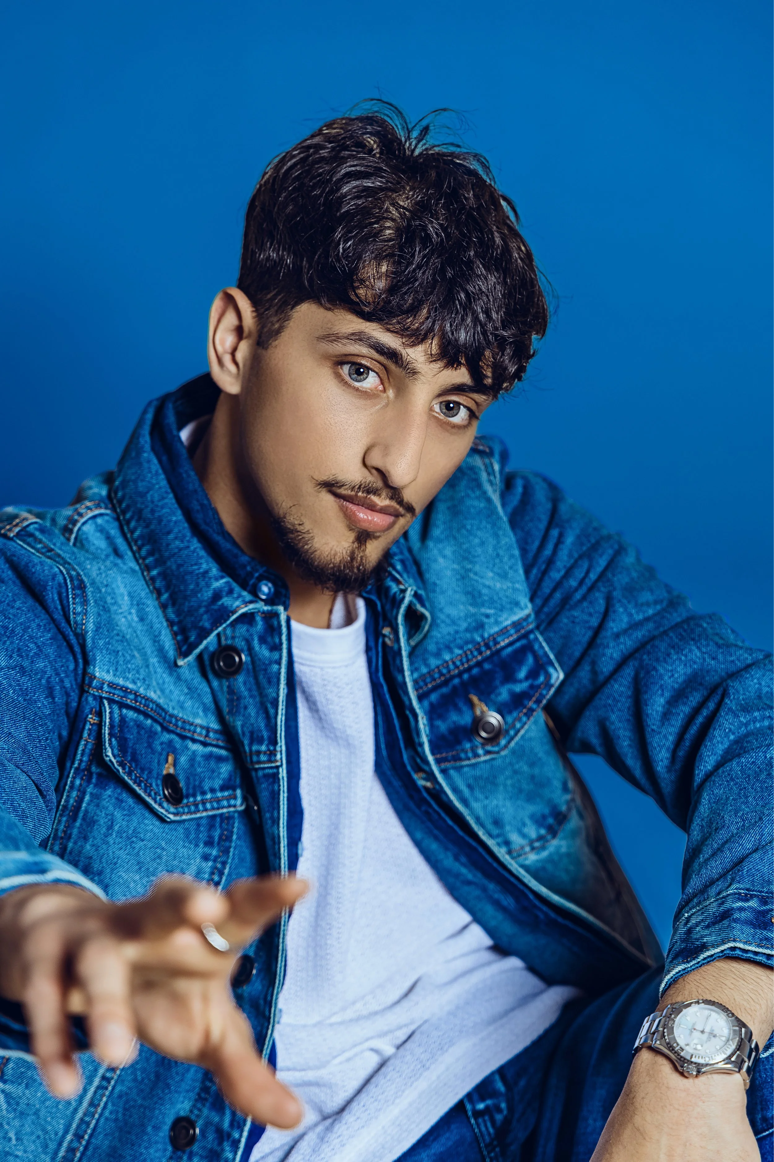 A young man with dark hair and blue eyes wearing a denim jacket, white t-shirt, and watch, sitting against a blue background and reaching towards the camera.