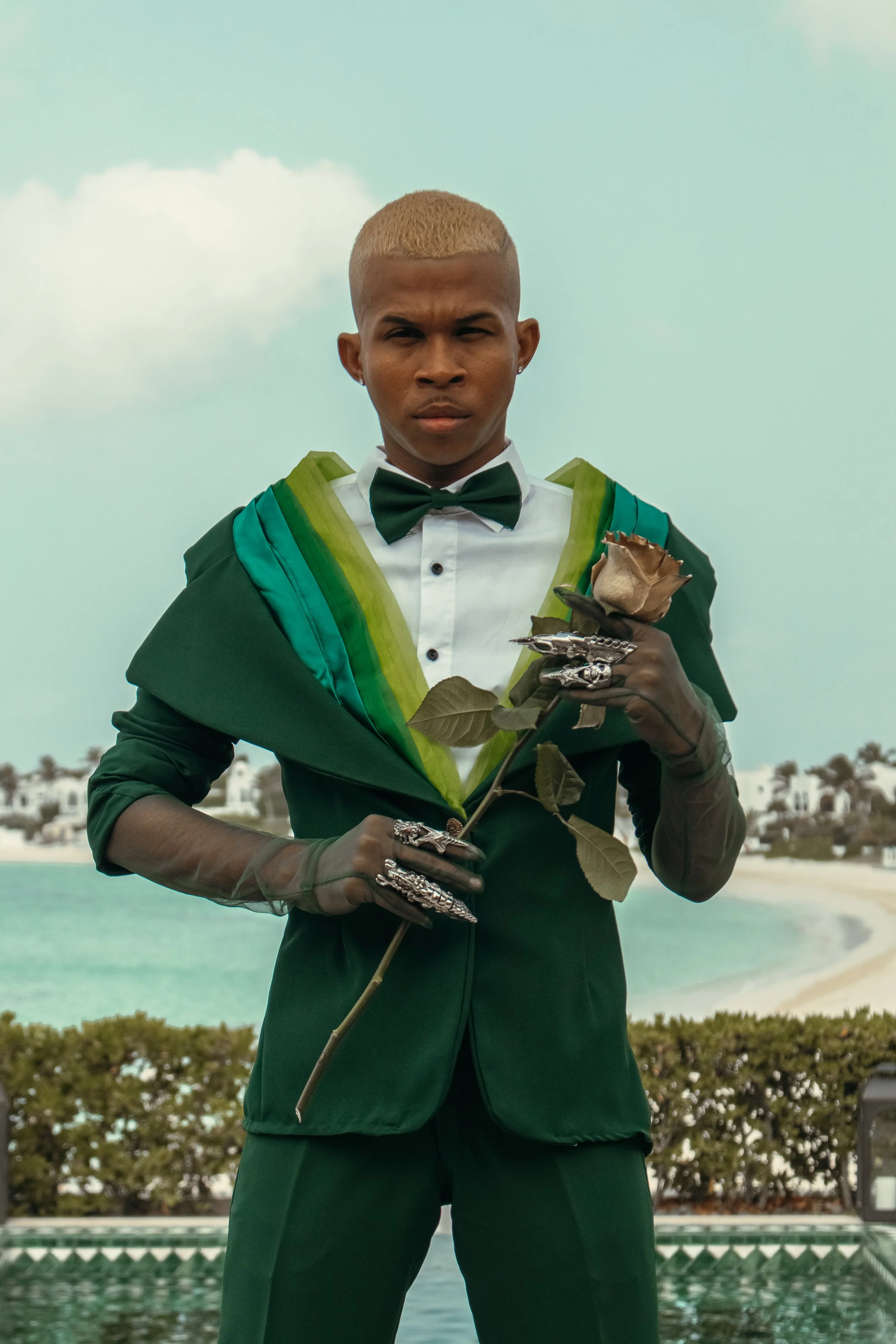 A person dressed in a dark green suit with a white shirt, black bow tie, and matching green scarf, holding a single rose. They are wearing decorative rings and gloves, standing outdoors with a beach and water in the background.