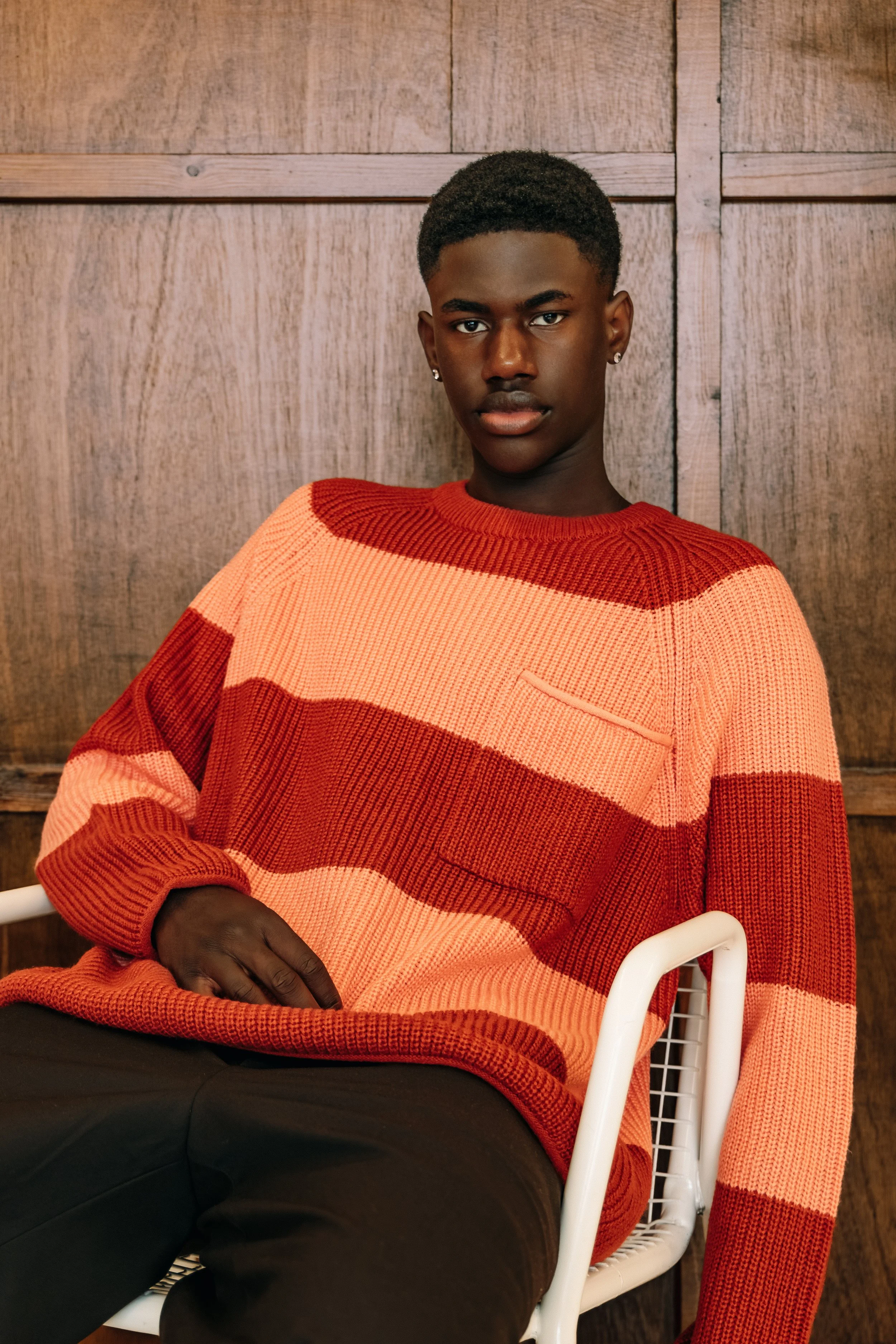 A young man with dark skin, short curly hair, wearing a red and orange striped knitted sweater, sitting in a white chair, against a wooden wall background.