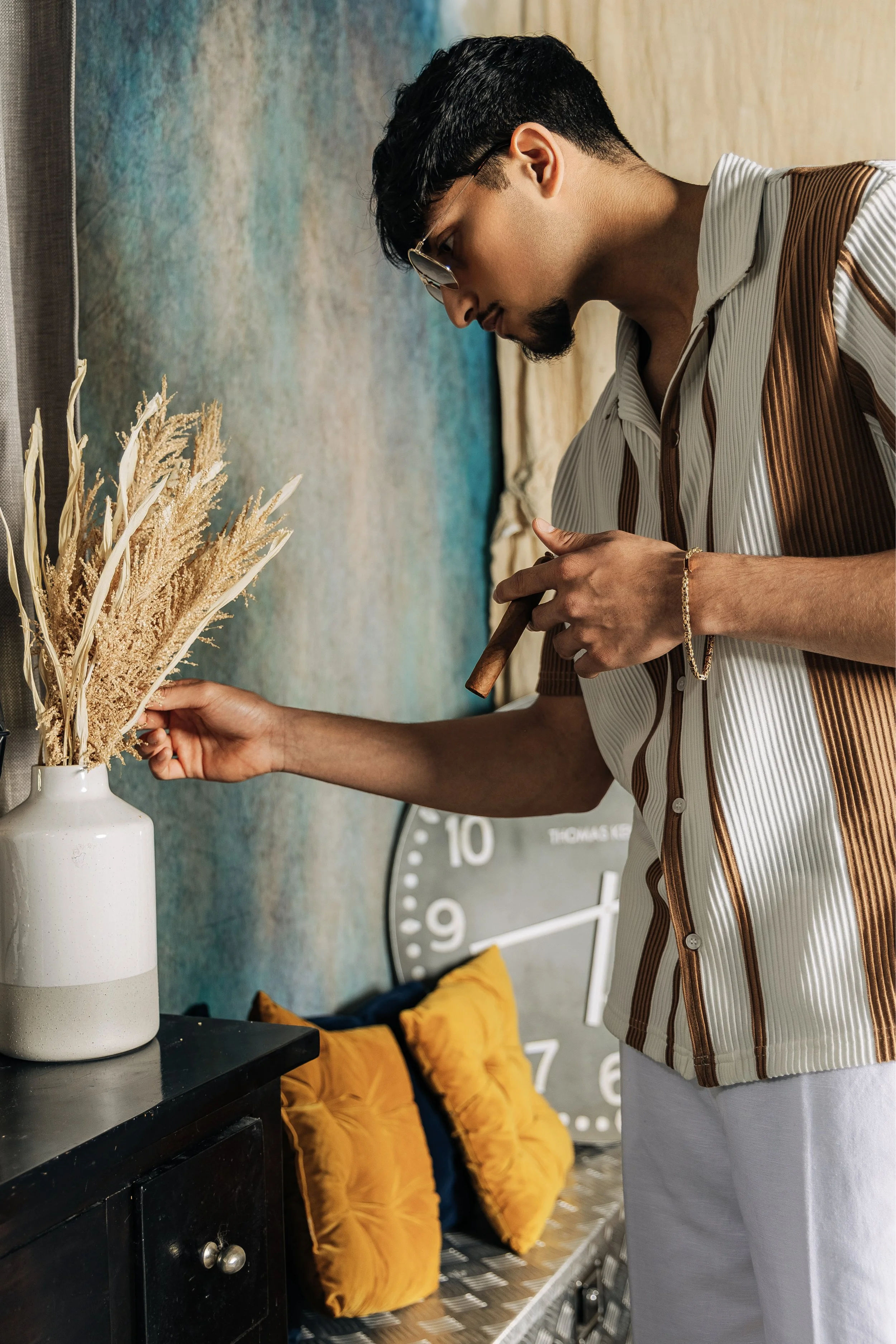 A man wearing glasses and a striped shirt arranging dried plants in a white vase on a black table, with a large wall clock and yellow cushions in the background.