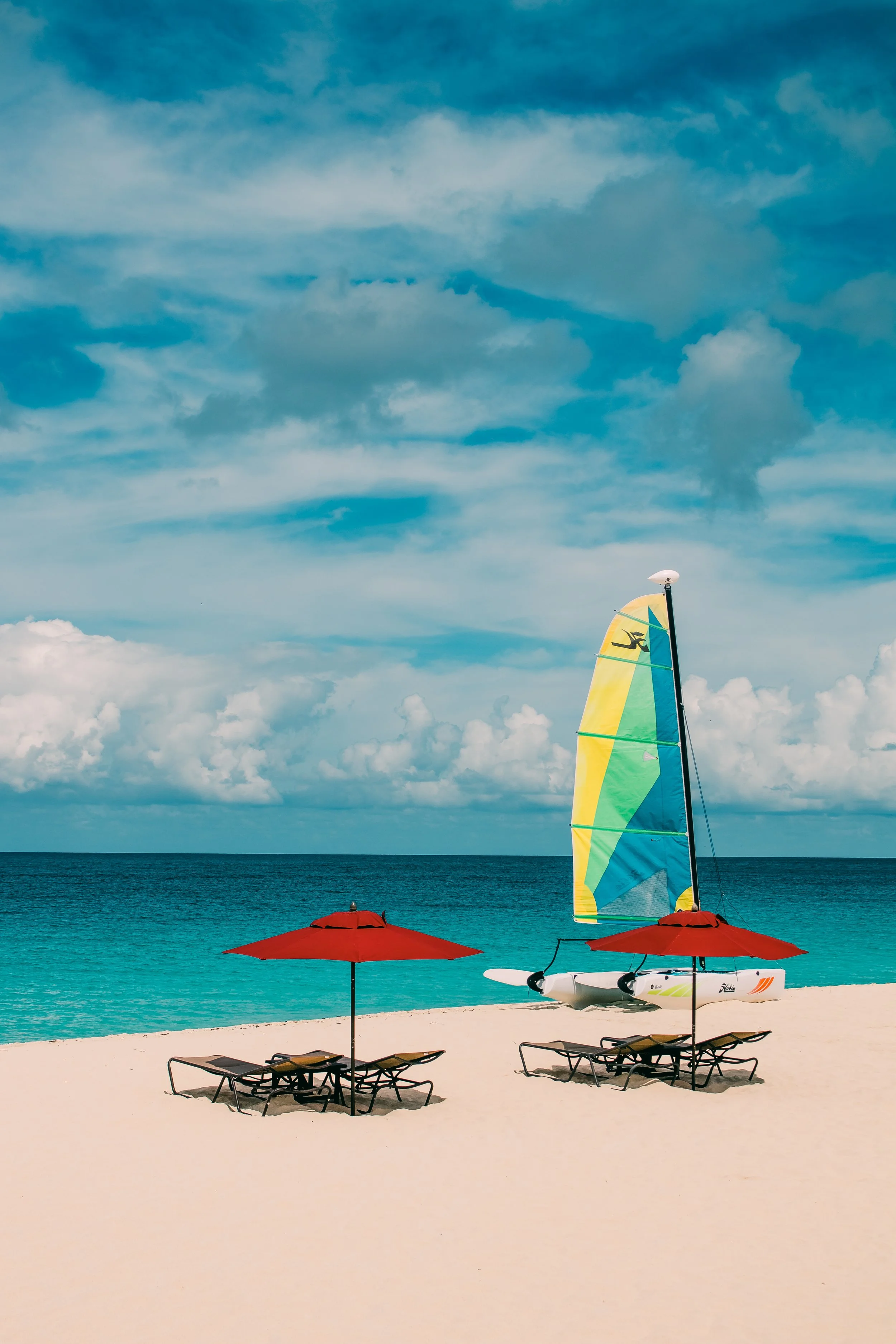 Beach scene with two red umbrellas, lounge chairs, sailboat with colorful sail, calm ocean, and partly cloudy sky.