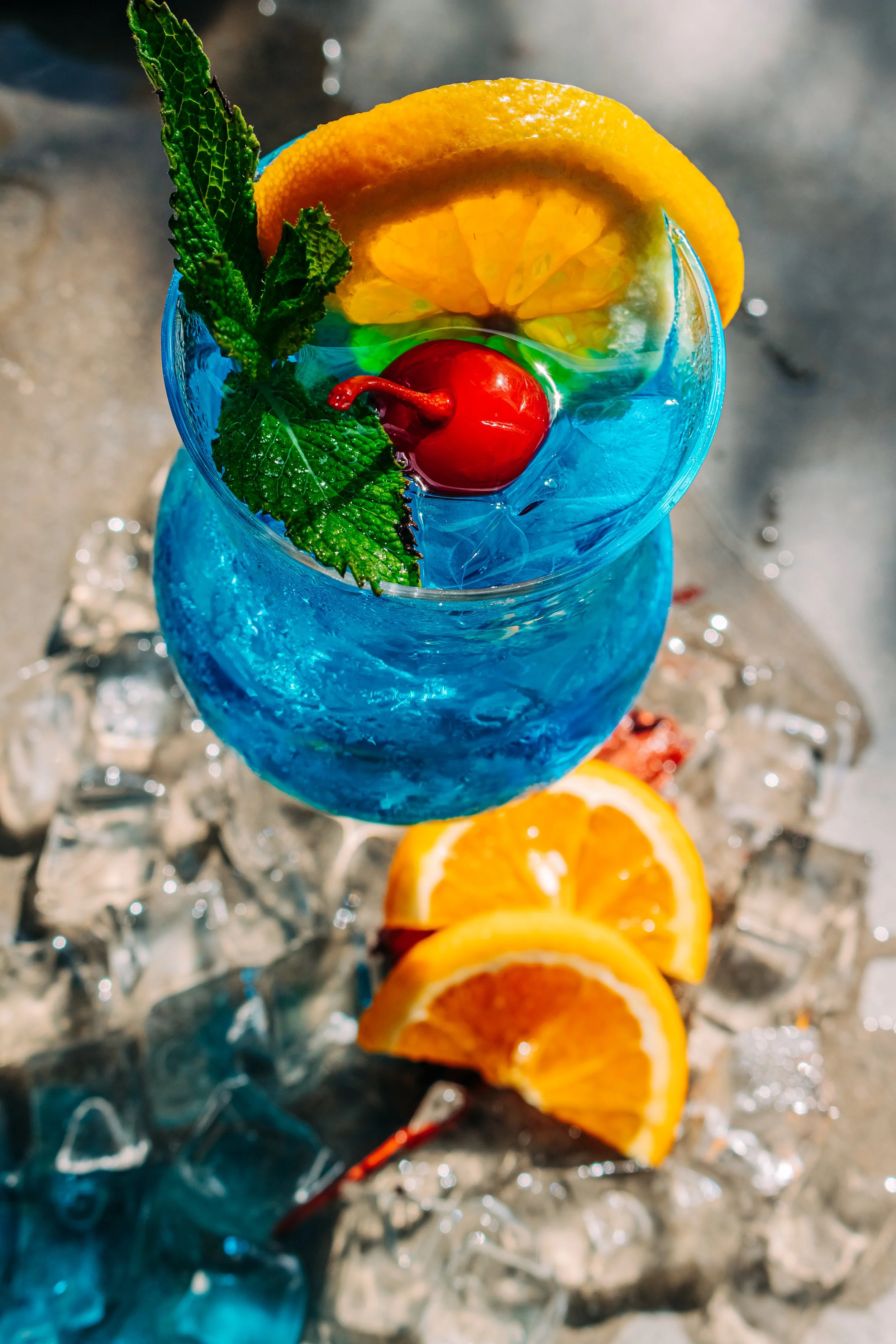 A tropical cocktail in a tall, blue glass garnished with a cherry, lemon wheel, and fresh mint leaves, with lemon slices on ice underneath.