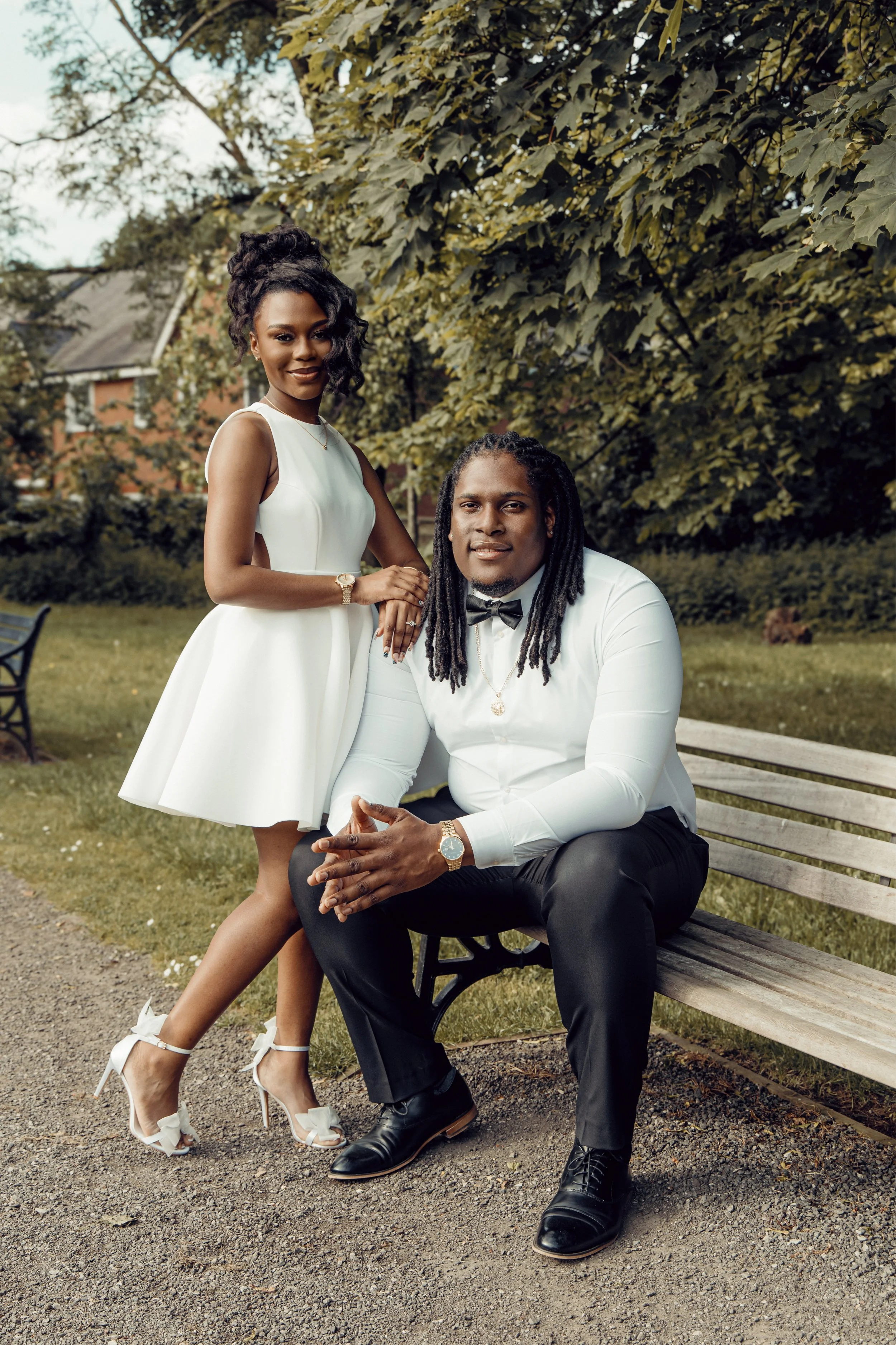 A stylishly dressed couple outdoors sitting on a park bench, with the woman standing beside the man. They are both smiling and looking at the camera, surrounded by trees and greenery.