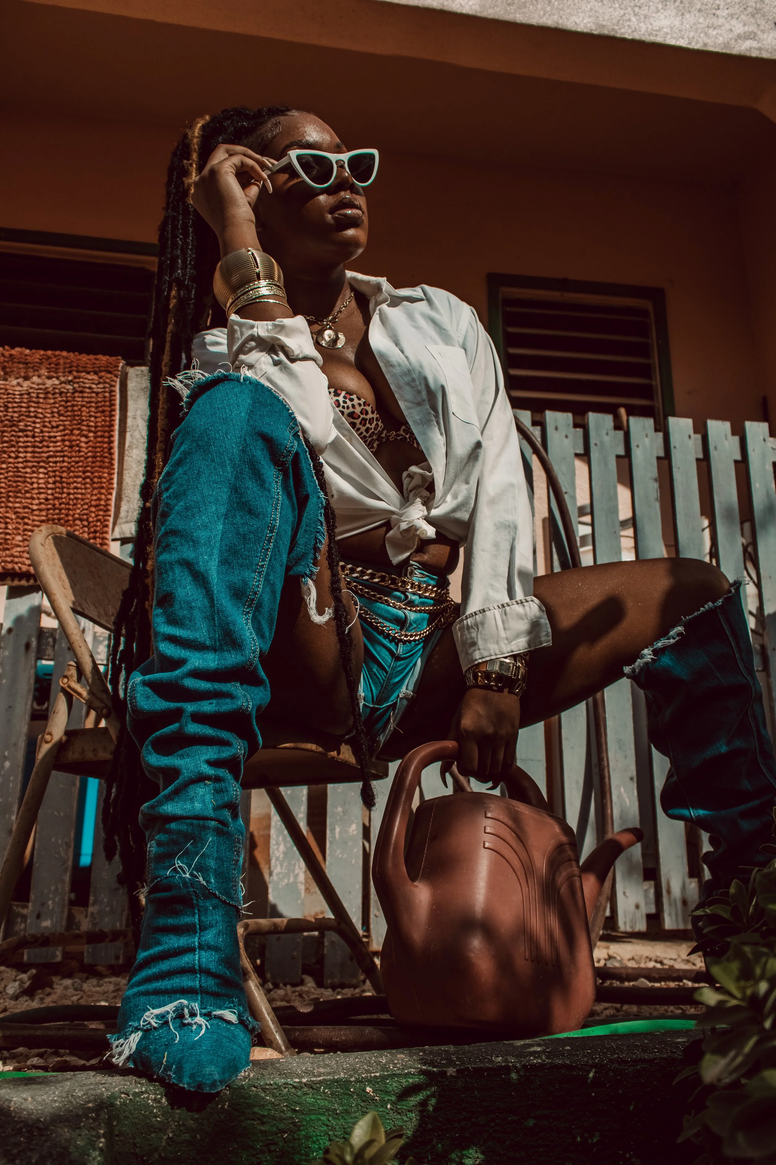 A woman with dreadlocks wearing sunglasses, a white shirt, denim shorts, and jewelry, sitting on a chair with one leg up, holding a watering can, with a dark background and plants in the foreground.