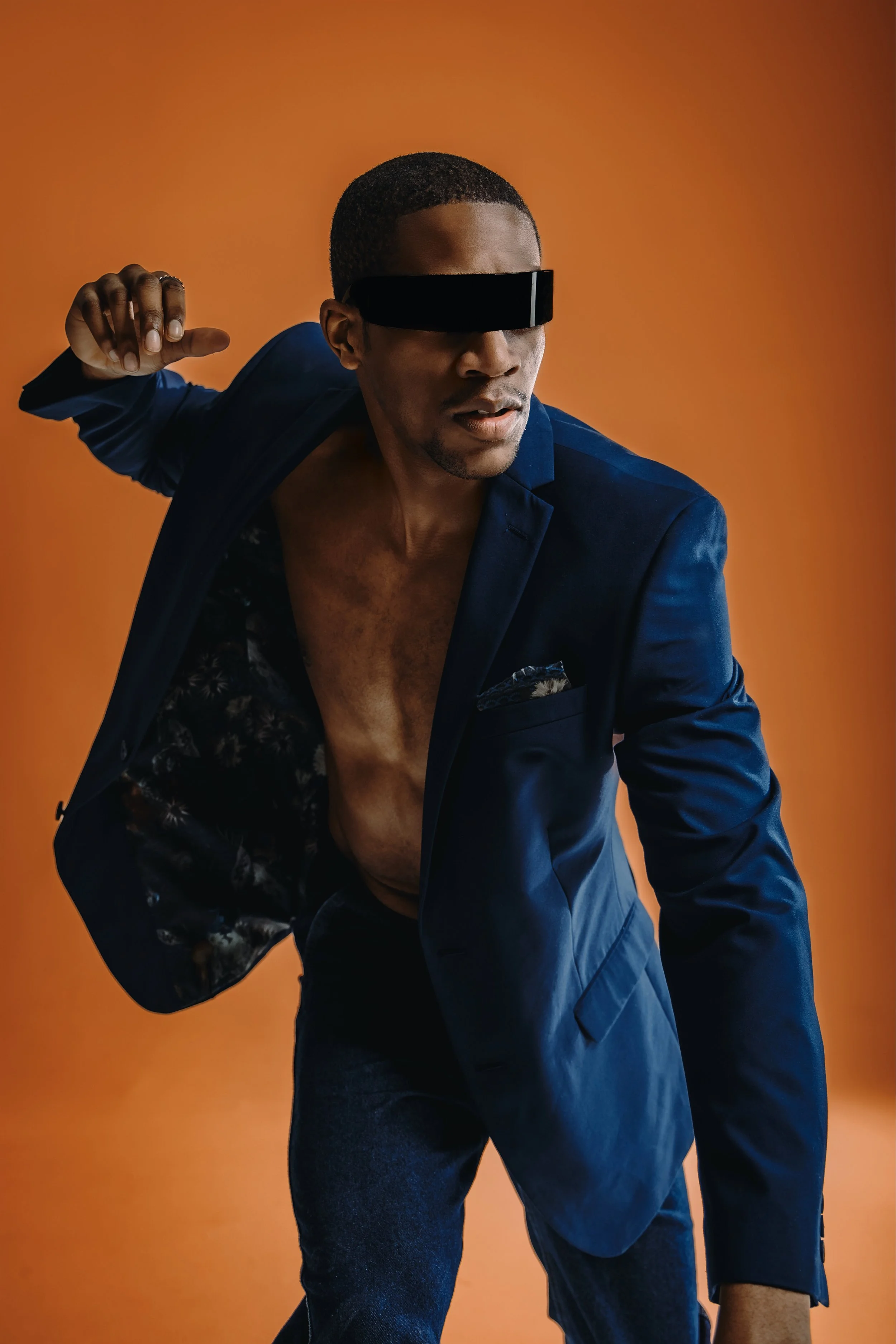 A man with dark skin dressed in a blue suit with the shirt unbuttoned, wearing black futuristic sunglasses, posing against an orange background.