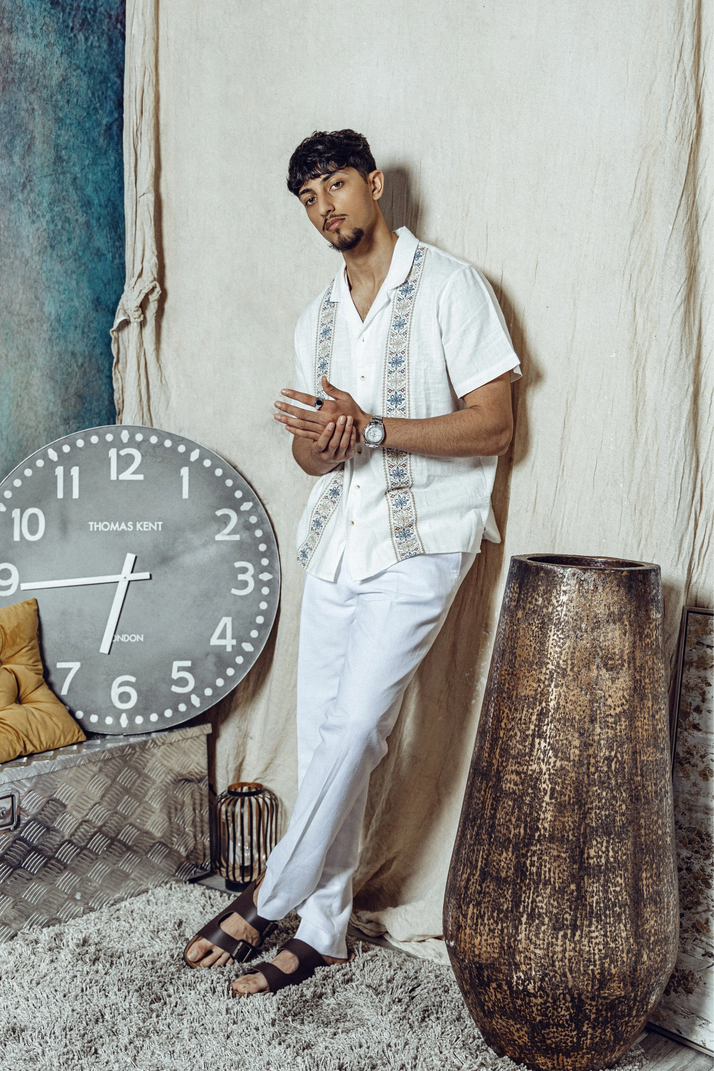 A young man with curly hair and a beard, dressed in white clothing, leaning against a beige fabric backdrop. There is a large round gray clock with white numbers and hands on a side table, and a tall, textured brown vase nearby. A pillow and framed a