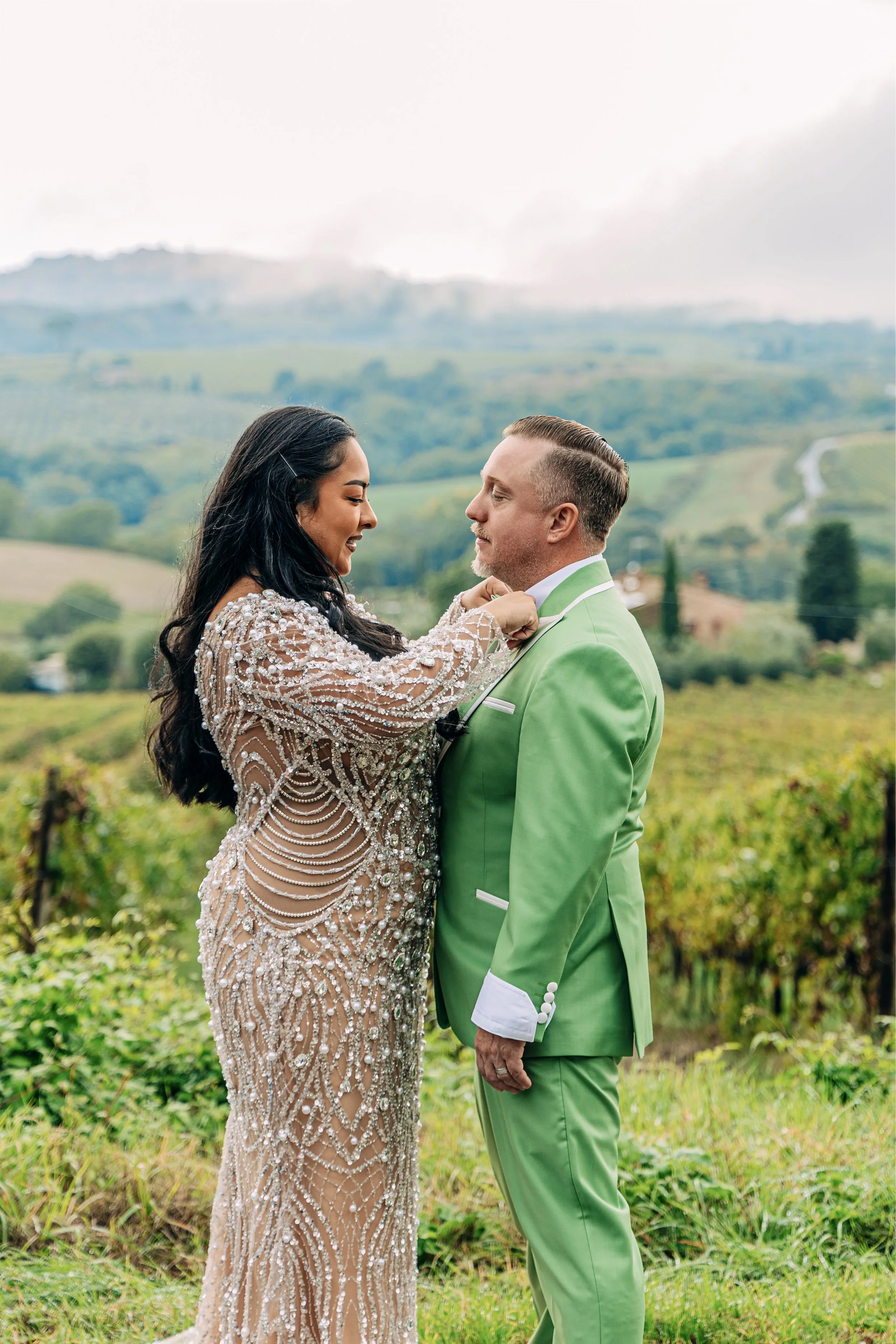A woman in a sparkling, intricate gown is holding a man's face in her hands as they smile at each other in an outdoor vineyard setting with rolling hills in the background.