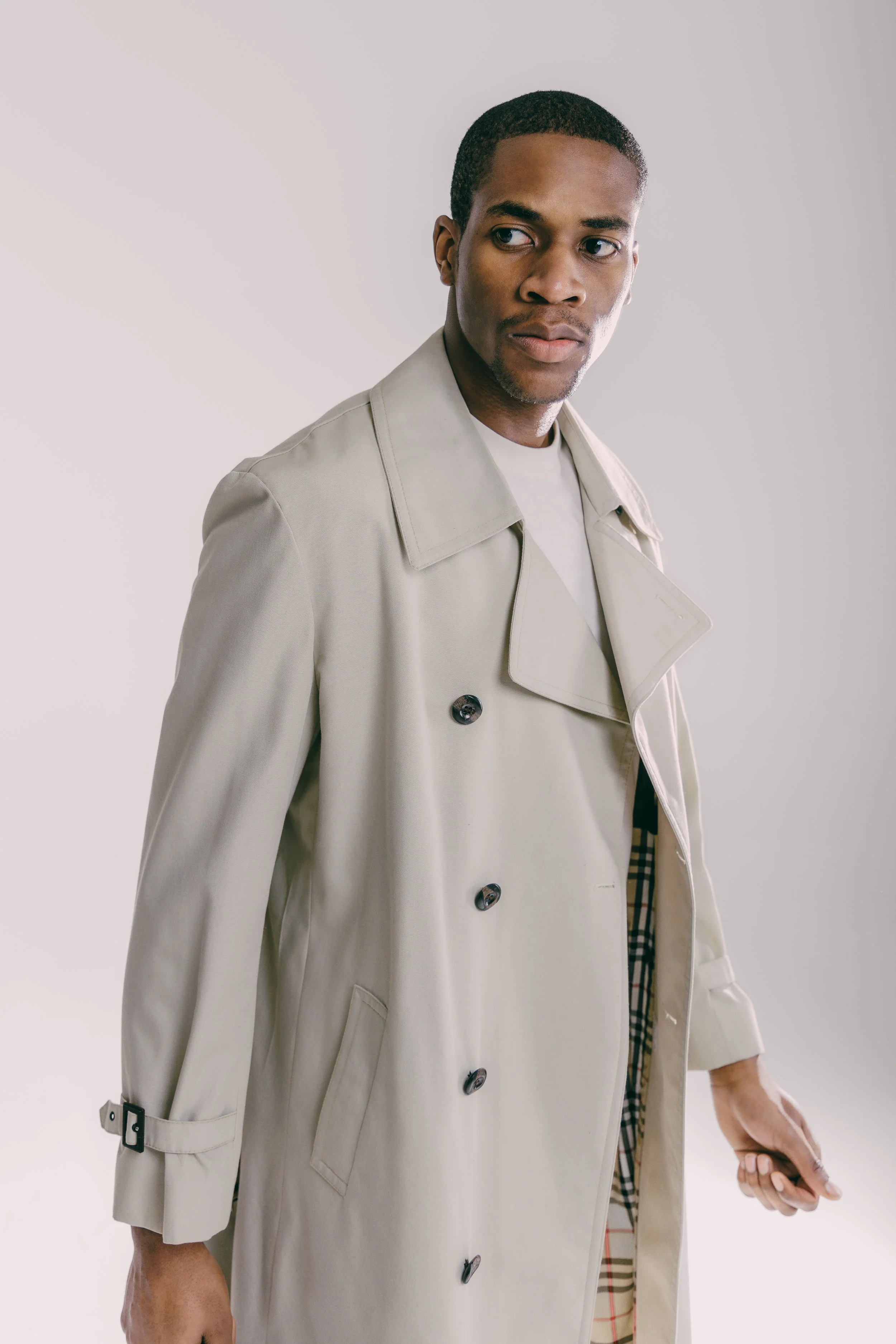 A man wearing a beige trench coat with dark buttons, standing against a plain background, looking to his left.