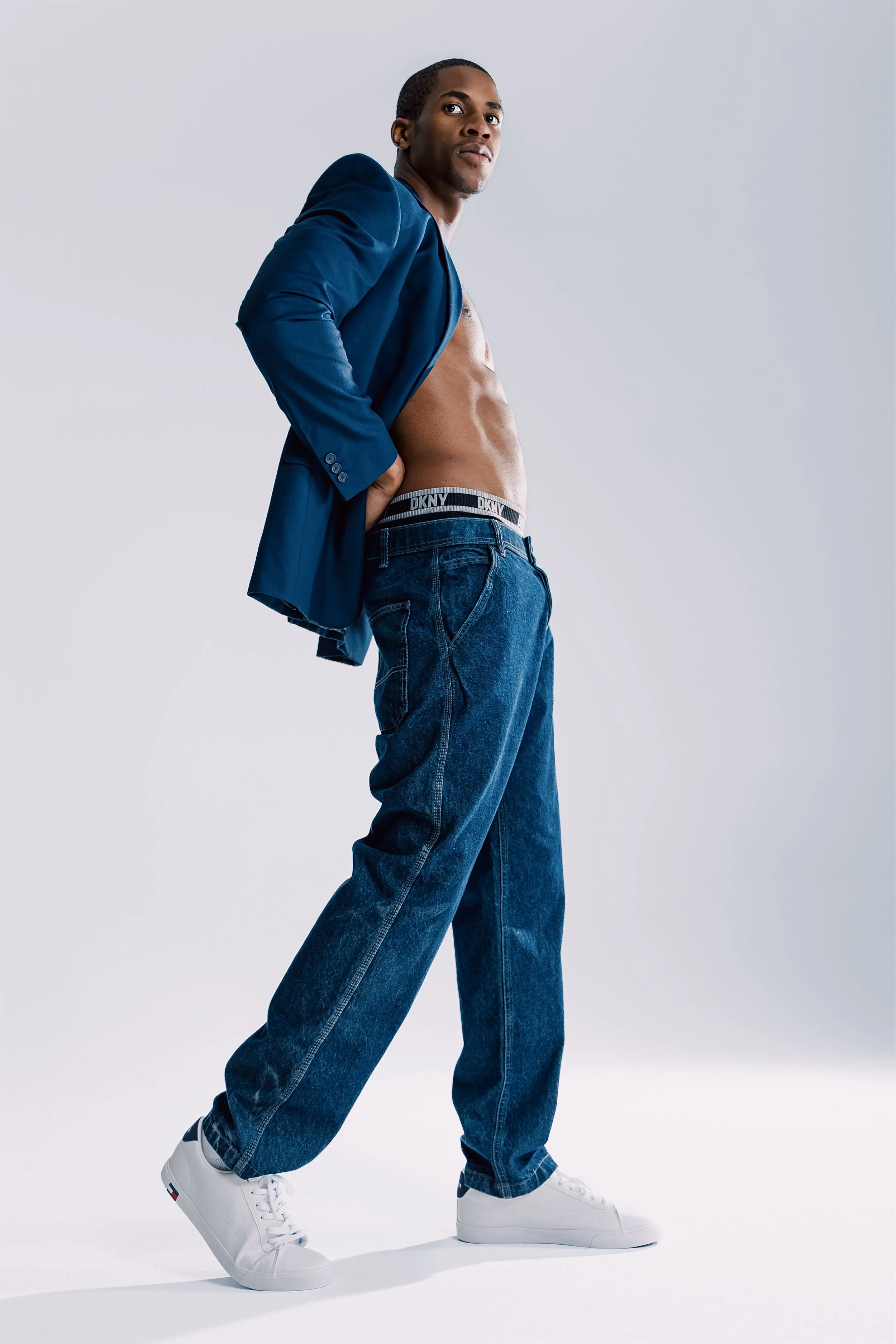 A young man with a muscular build posing confidently against a plain light gray background, wearing dark blue high-waisted jeans, a partially unbuttoned blue blazer, and white sneakers. His underwear with a DKNY waistband is visible as he adjusts his