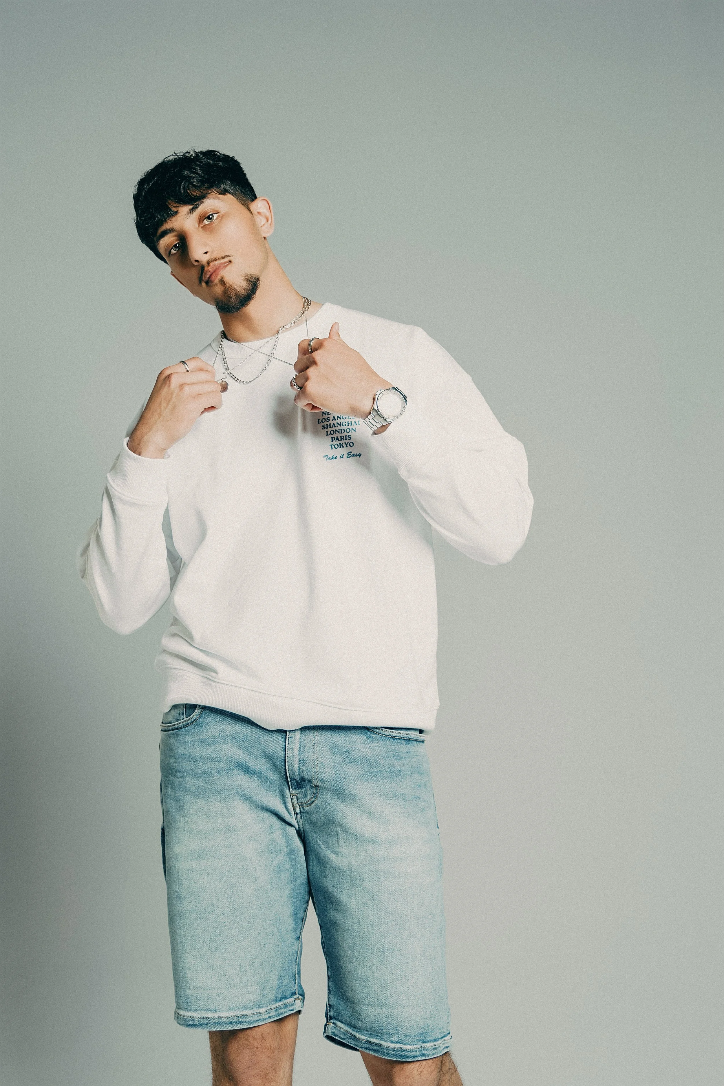 Young man with dark, wavy hair and facial hair, wearing a white sweatshirt and light blue denim shorts, standing against a plain light gray background, holding jewelry around his neck.