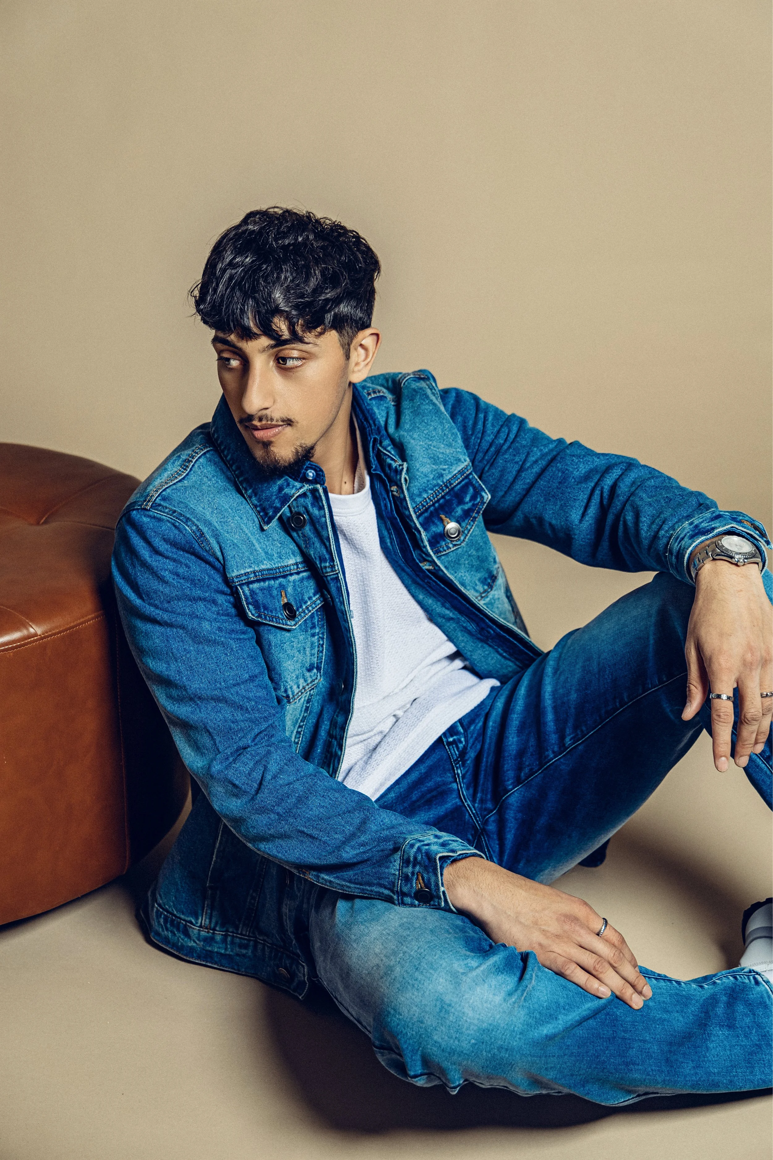 A young man with dark hair and facial hair sitting on the floor, leaning against a brown sofa, wearing a blue denim jacket and jeans, white t-shirt, and a watch, looking to his left.