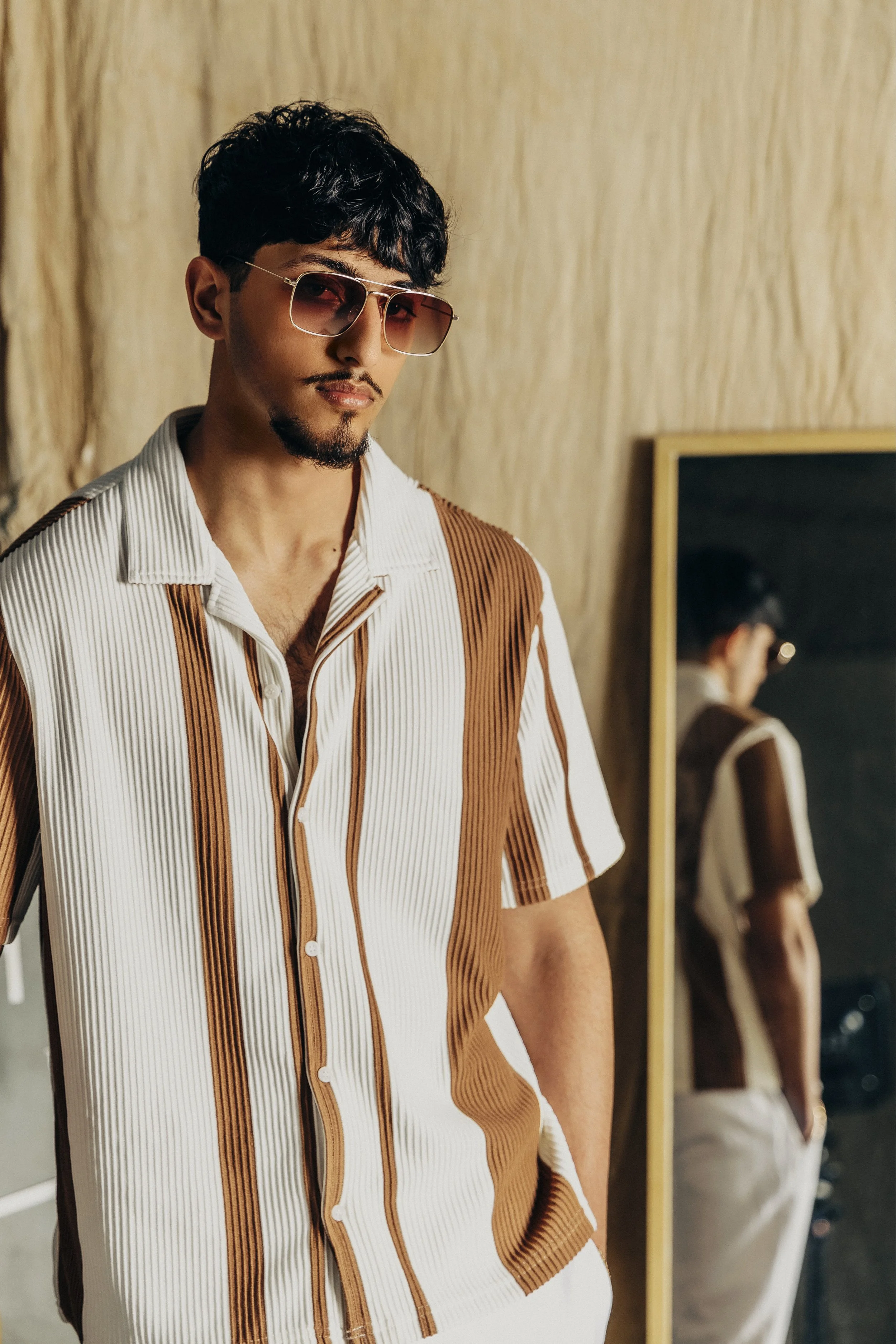 A man wearing sunglasses and a retro-style, short-sleeved, striped button-up shirt standing in front of a mirror.