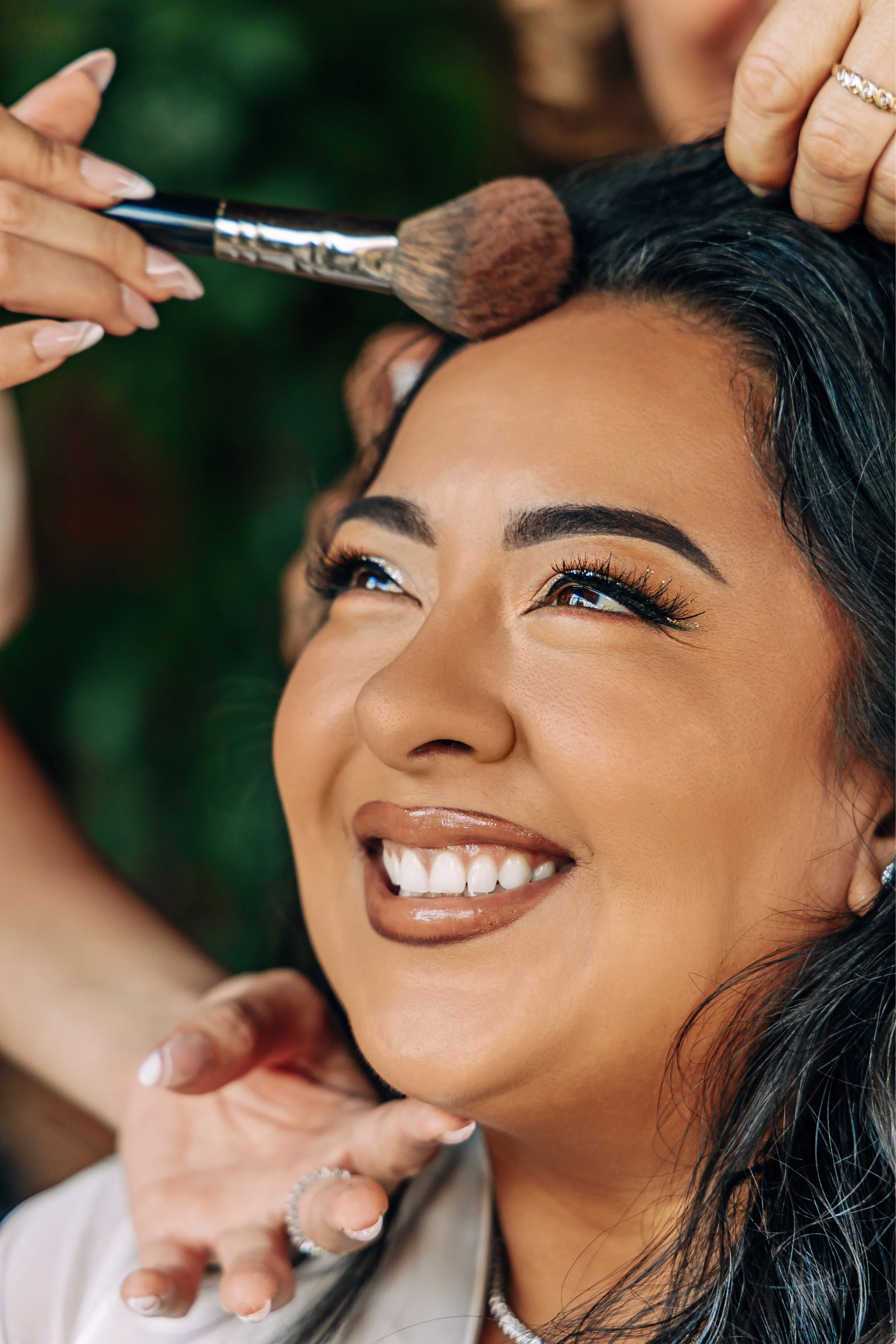 A woman smiling as she has her makeup applied by a makeup artist. The makeup artist is using a large brush on the woman's forehead. The woman has dark hair, defined eyebrows, and is smiling showing her teeth.