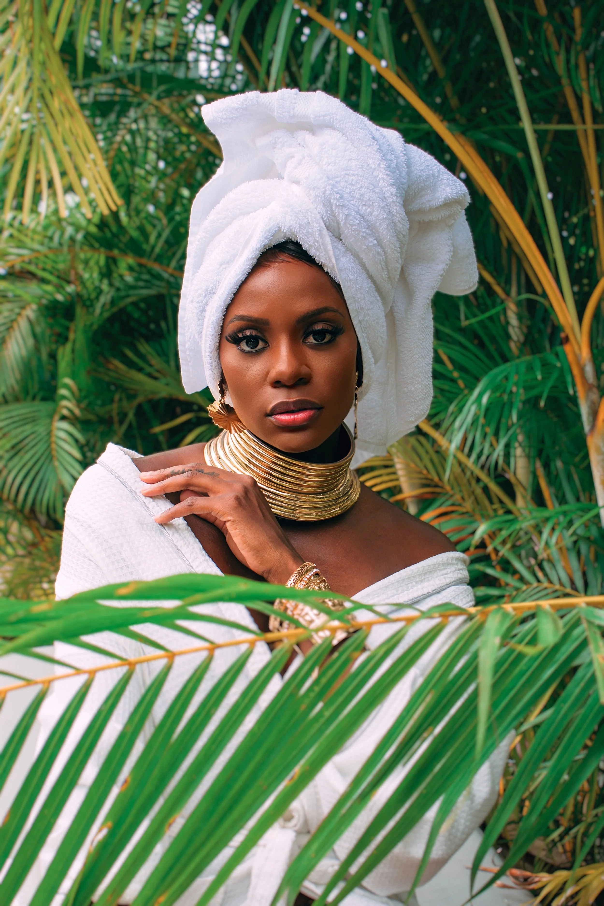 Portrait of a woman with dark skin, wearing a white towel wrapped around her head, gold jewelry, and a white garment, surrounded by green tropical plants.