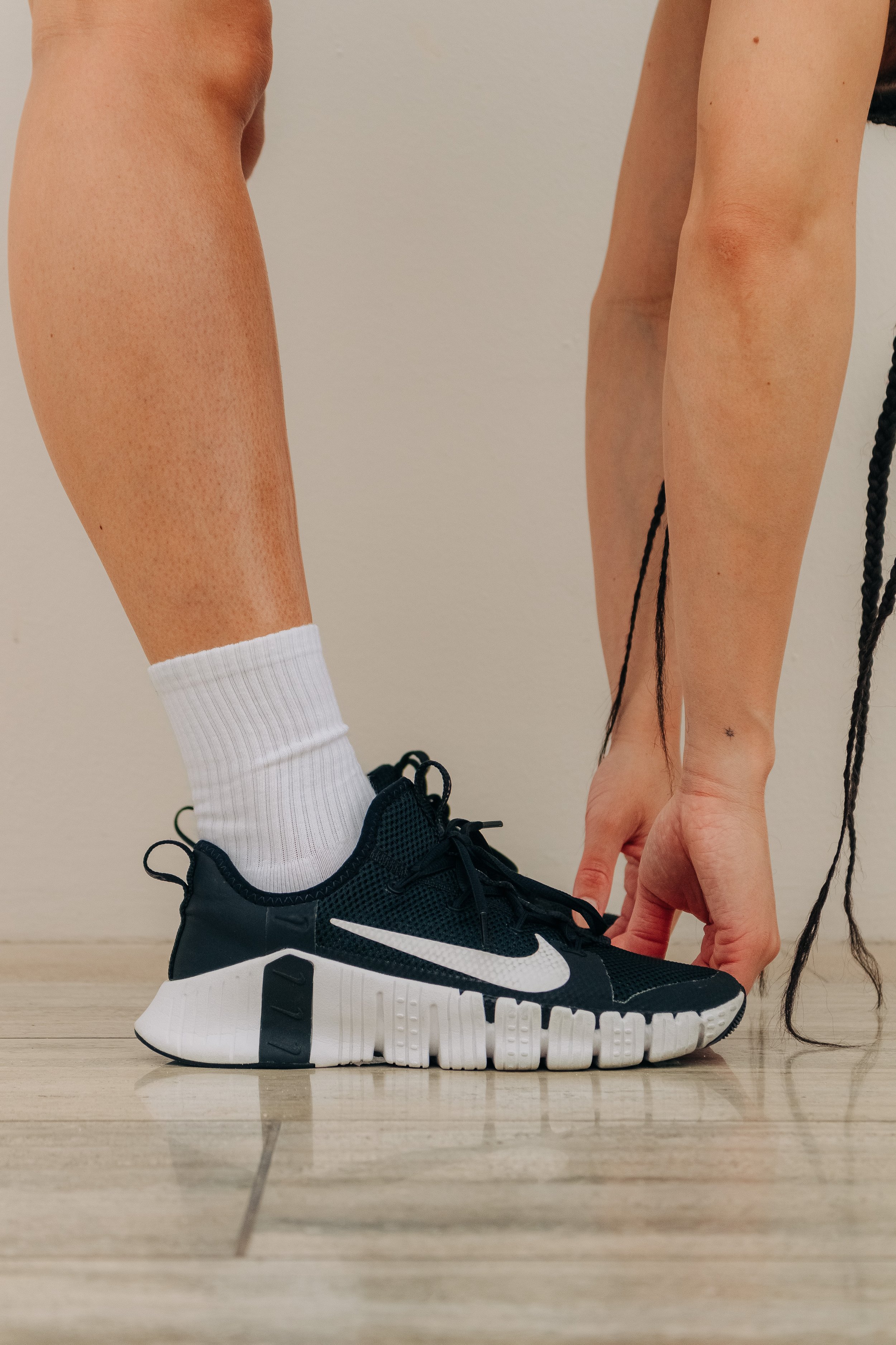 A person is tying the shoelaces of a black Nike athletic shoe with a white sole, while wearing white socks and squatting on a wooden floor.