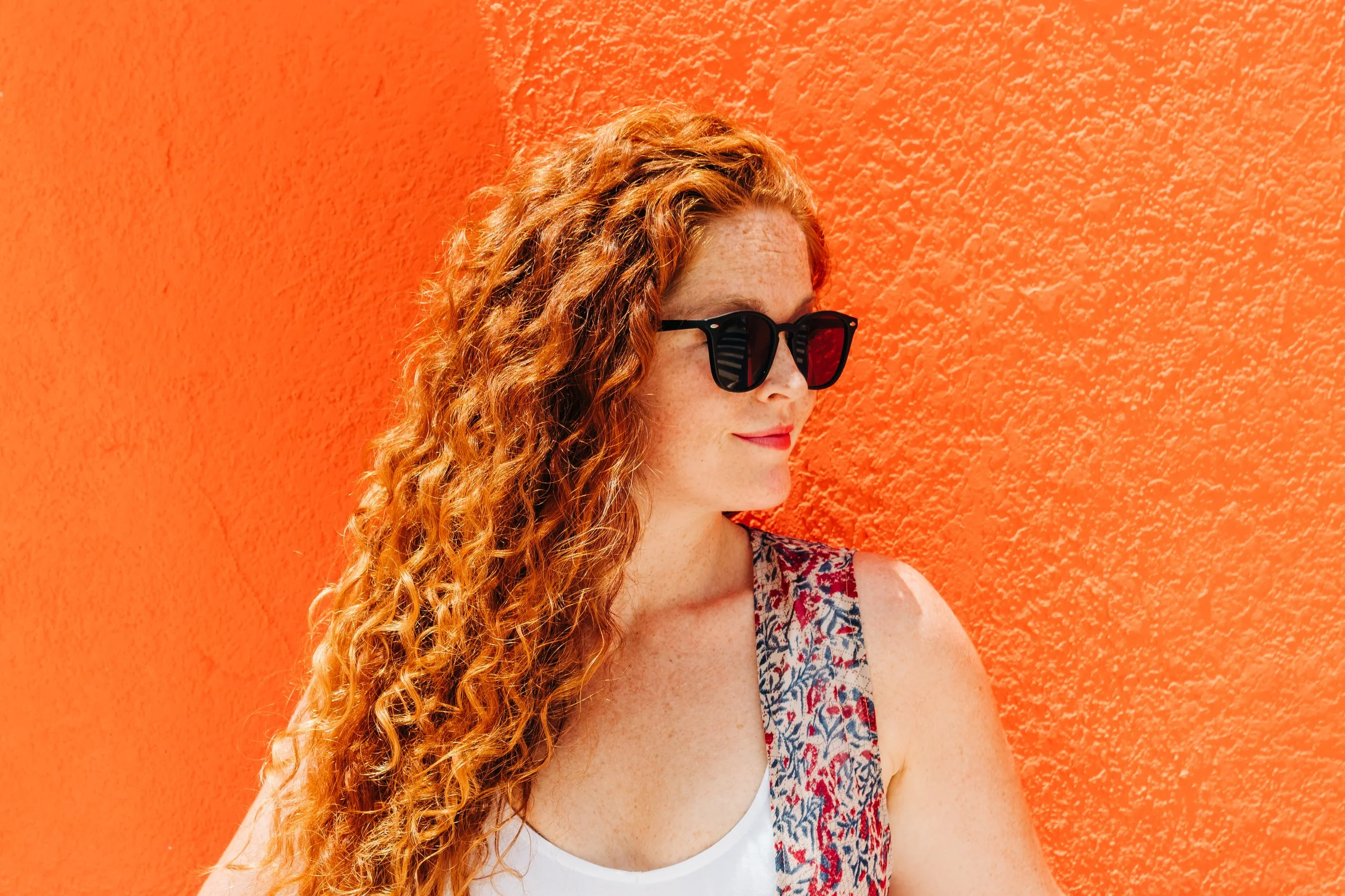 A woman with curly red hair and sunglasses standing in front of an orange textured wall.