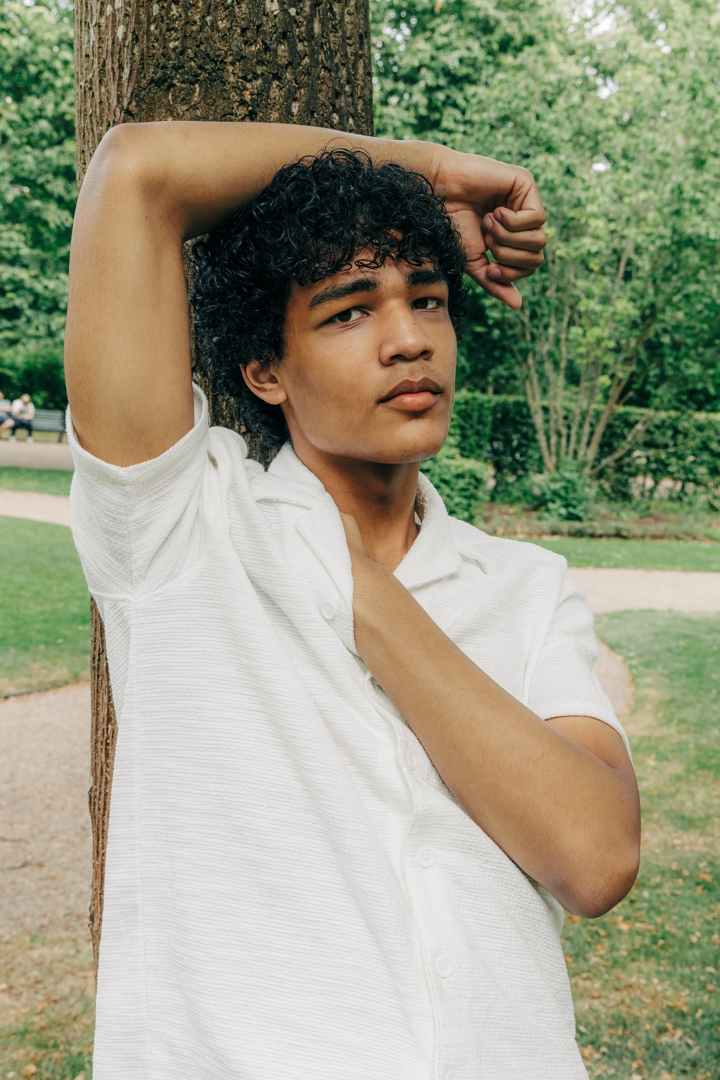 A young person with curly black hair and medium skin tone leaning against a tree in a park, wearing a white shirt, with a background of green trees and a park walkway.