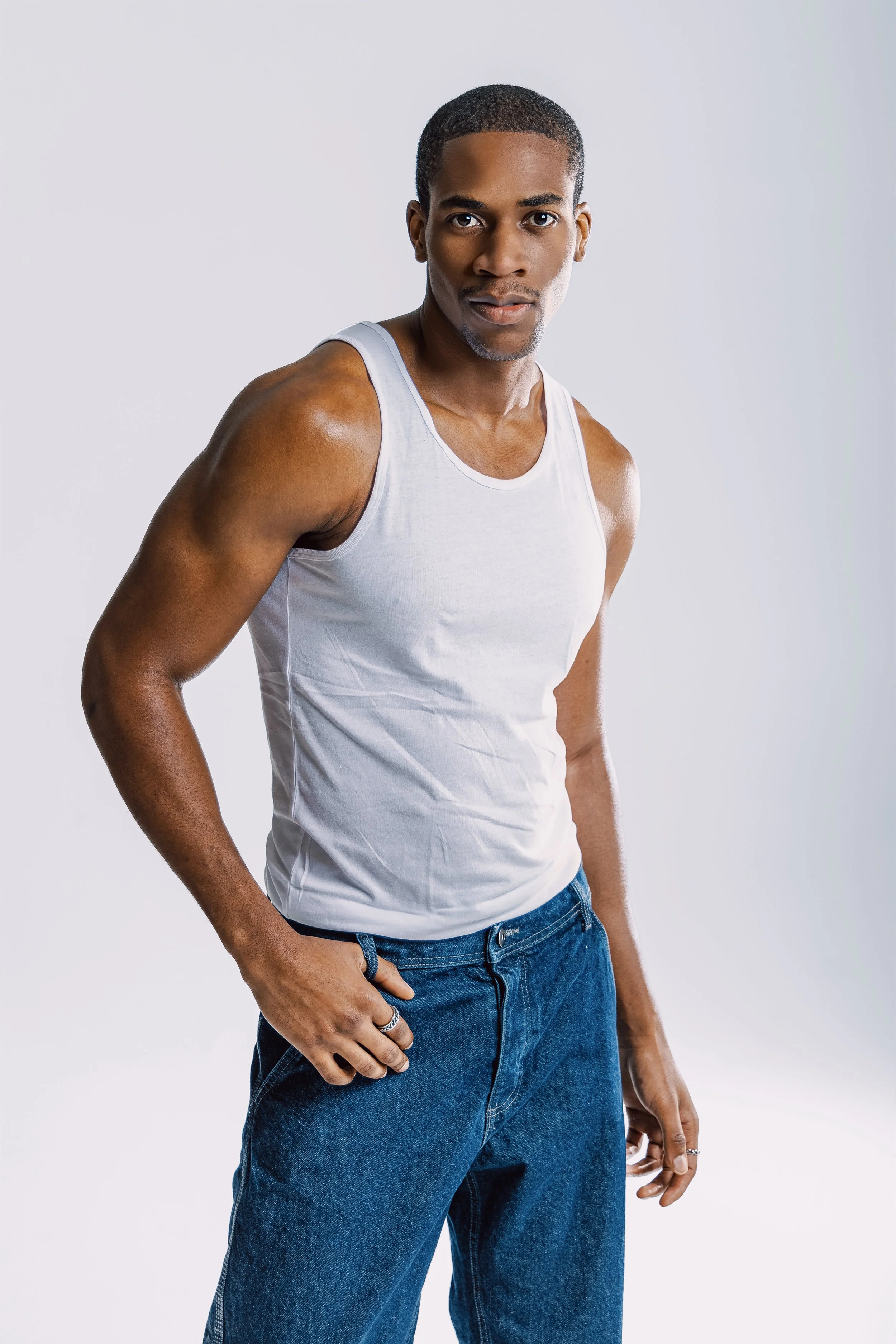 A young man with dark skin, wearing a white sleeveless tank top and blue jeans, standing against a plain light background.
