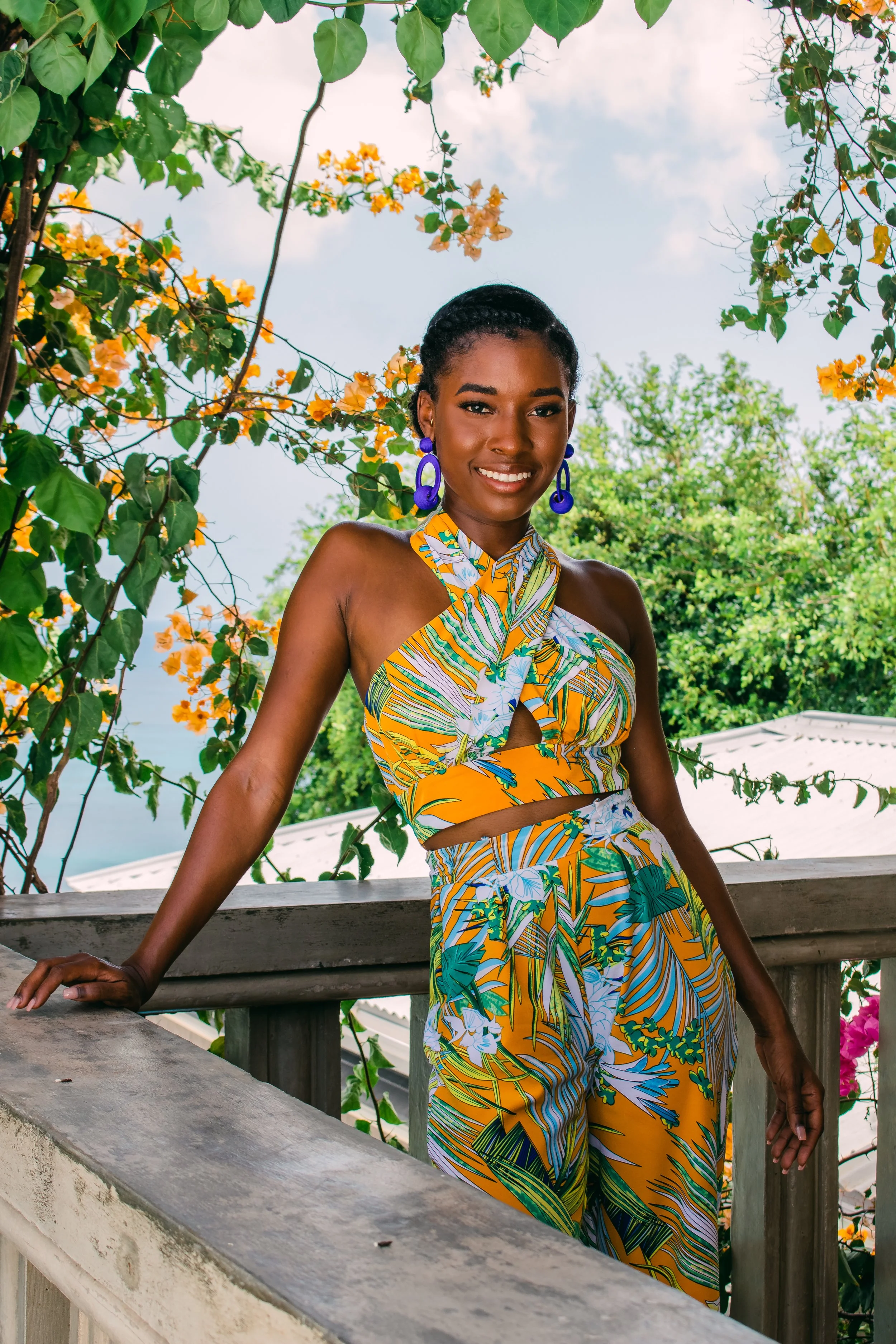 A woman standing outdoors on a balcony, wearing a colorful tropical print dress with a cutout, smiling, with green trees and orange flowers in the background.