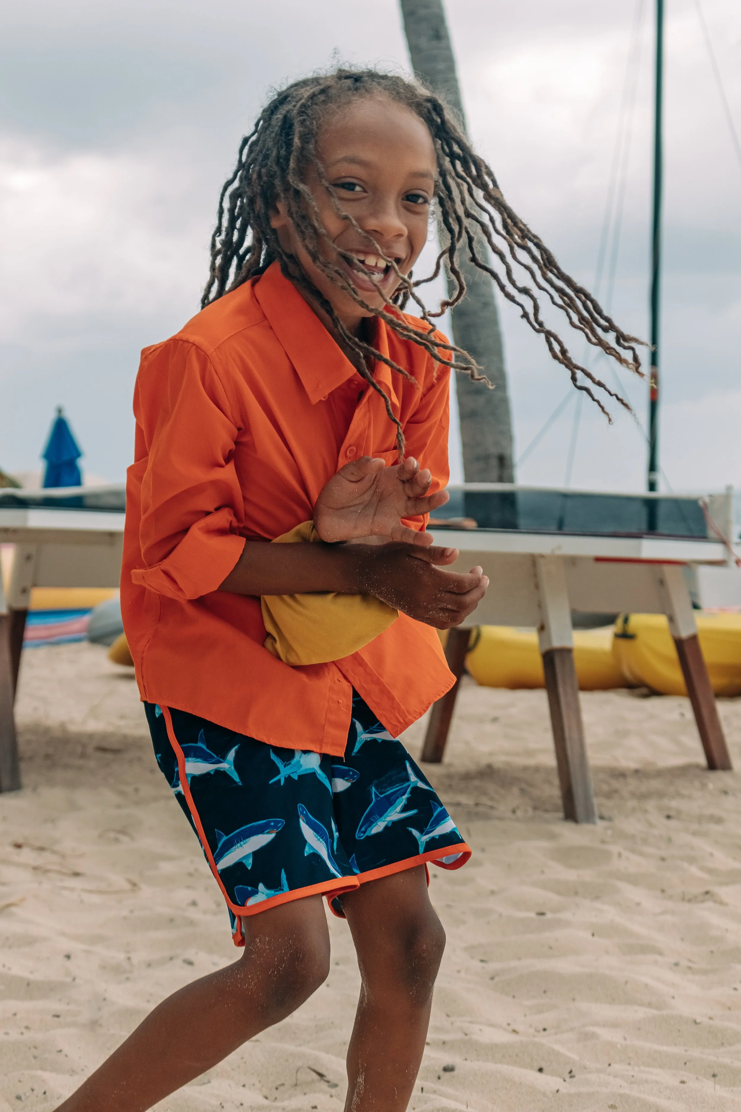 A young girl with dreadlocks smiling and playing on the beach, wearing an orange shirt and patterned shorts, with boats and a cloudy sky in the background.