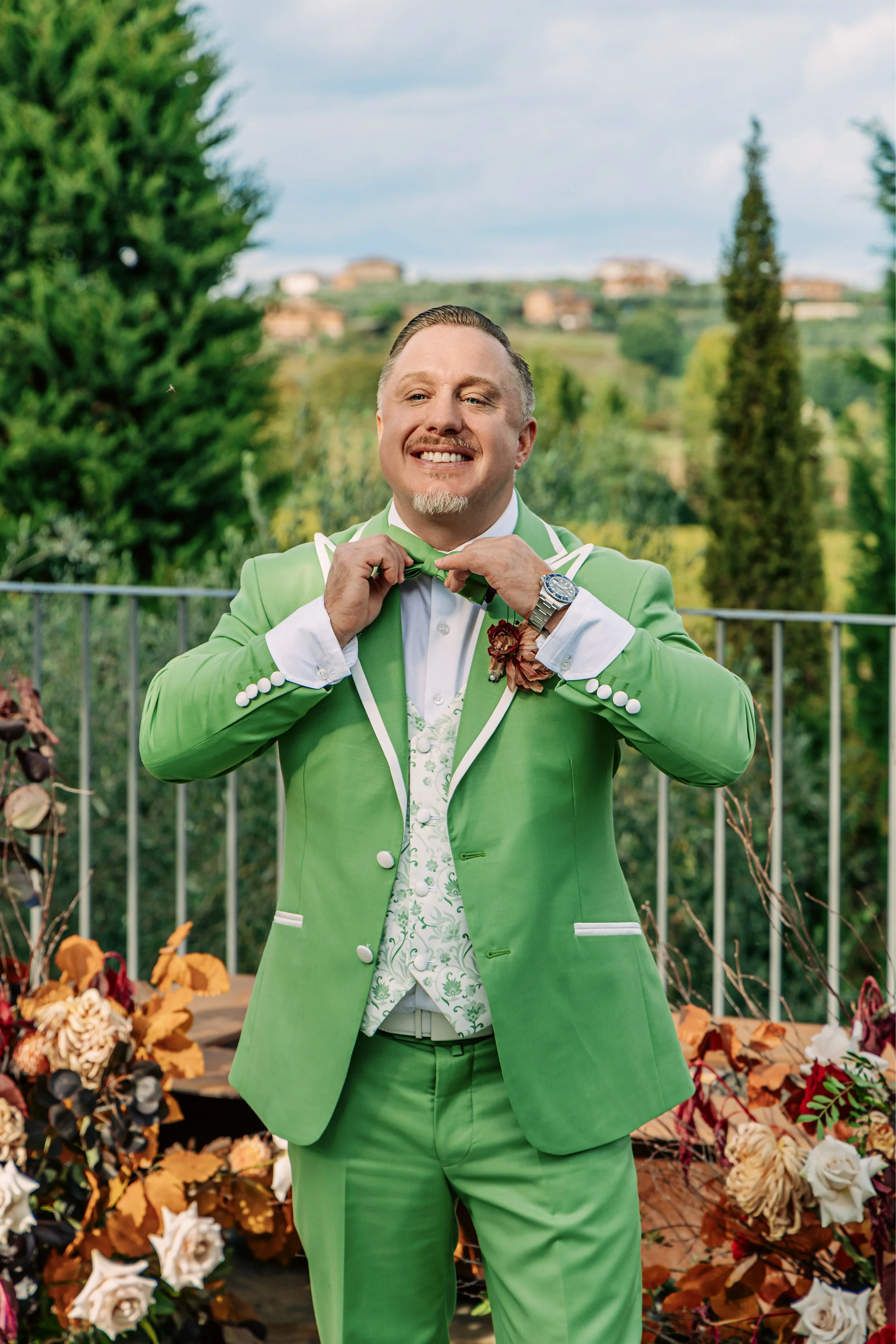 A man in a bright green suit adjusting his green bowtie outdoors, surrounded by autumn flowers and plants, with a scenic hillside in the background.